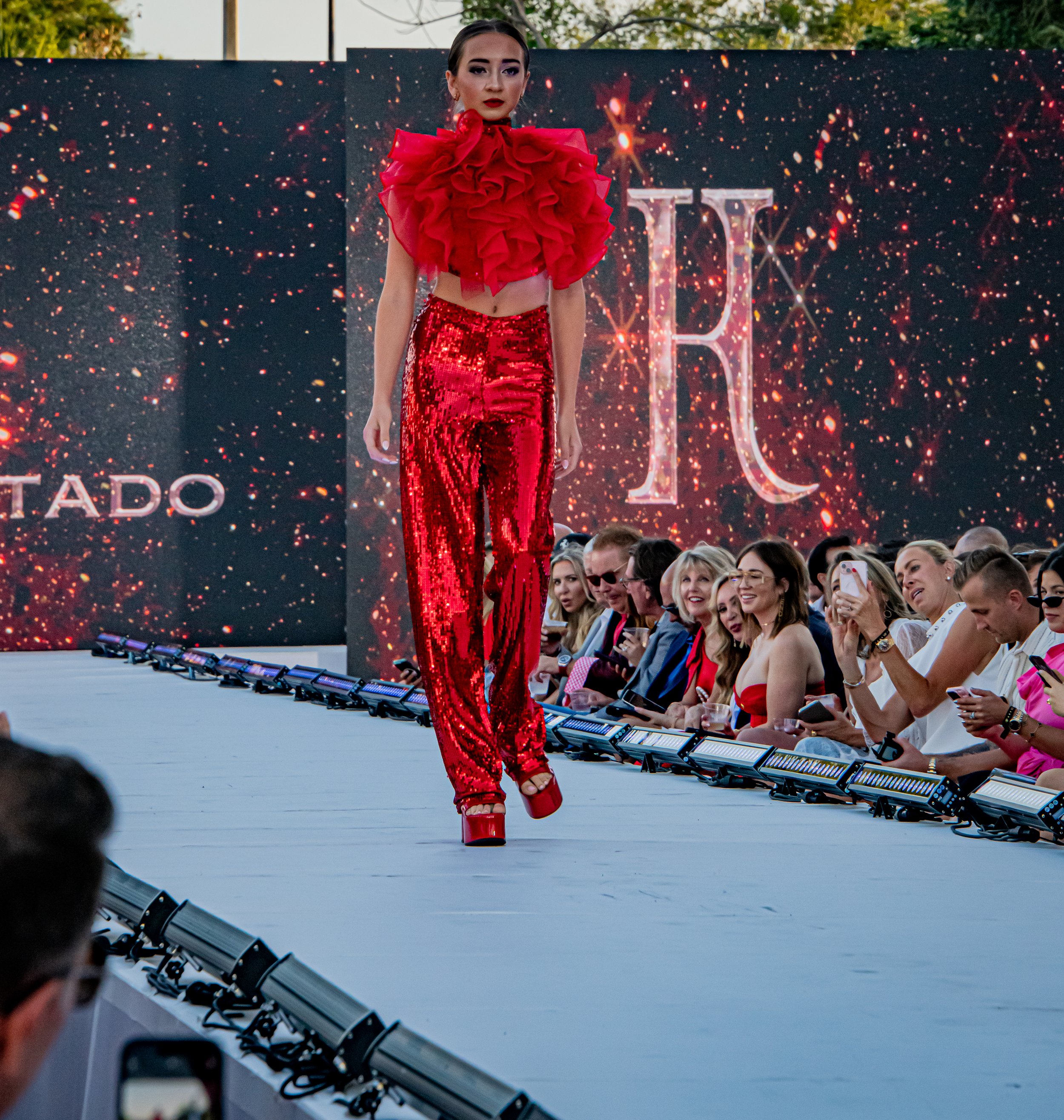 A model walking down the runway in a red, ruffled top and shiny, sequined red pants at a fashion show with an audience watching.