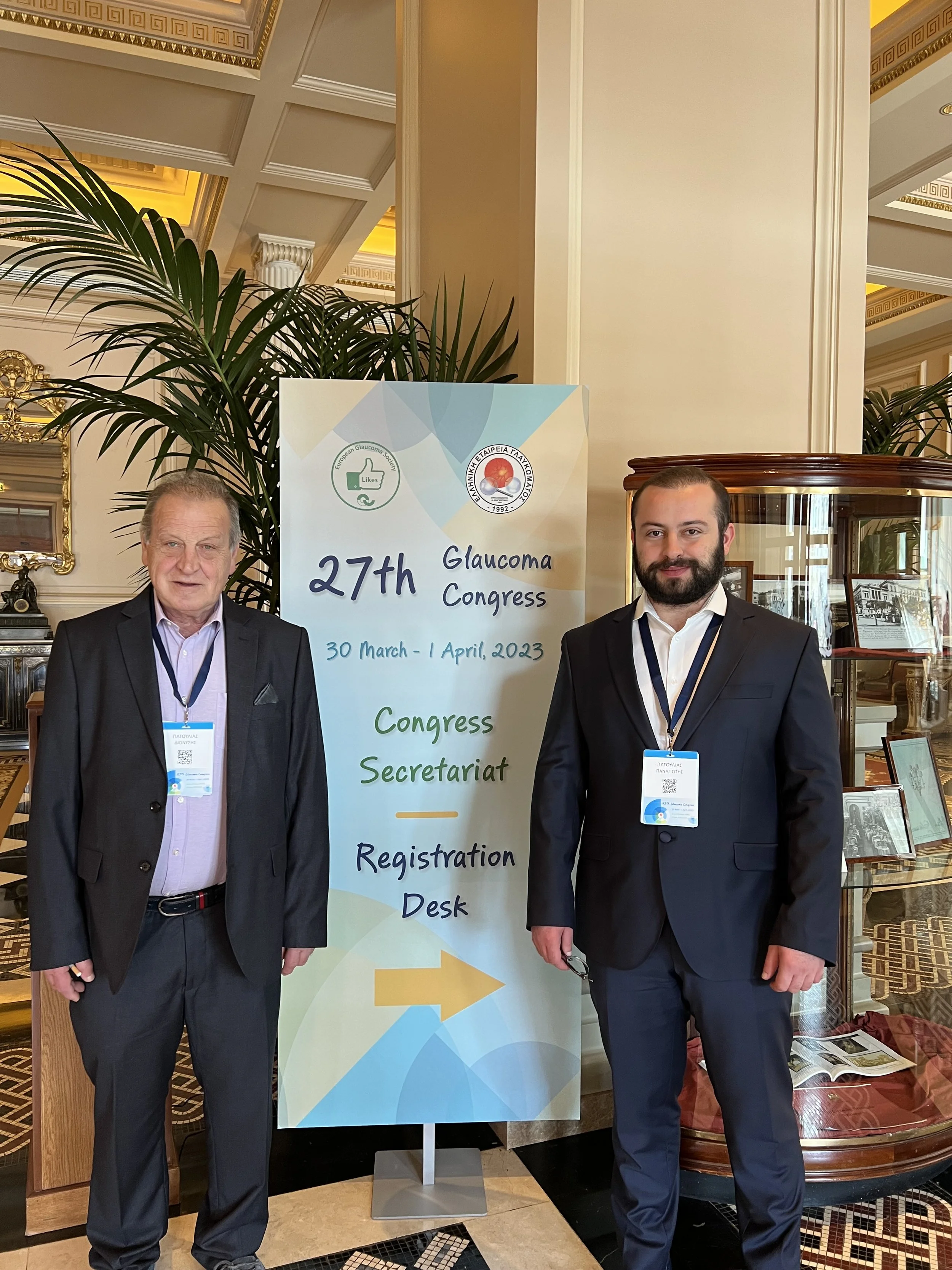 Two men in suits and conference badges standing next to a large sign at the 27th Glaucoma Congress, held from March 30 to April 1, 2023. The sign indicates registration desk and secretariat directions, with a decorative interior setting and plants in the background.