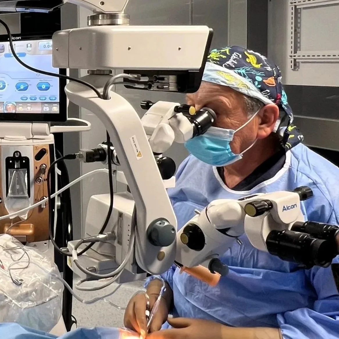 A surgeon wearing a surgical mask, cap, and gown looking through a microscope during an operation in an operating room.