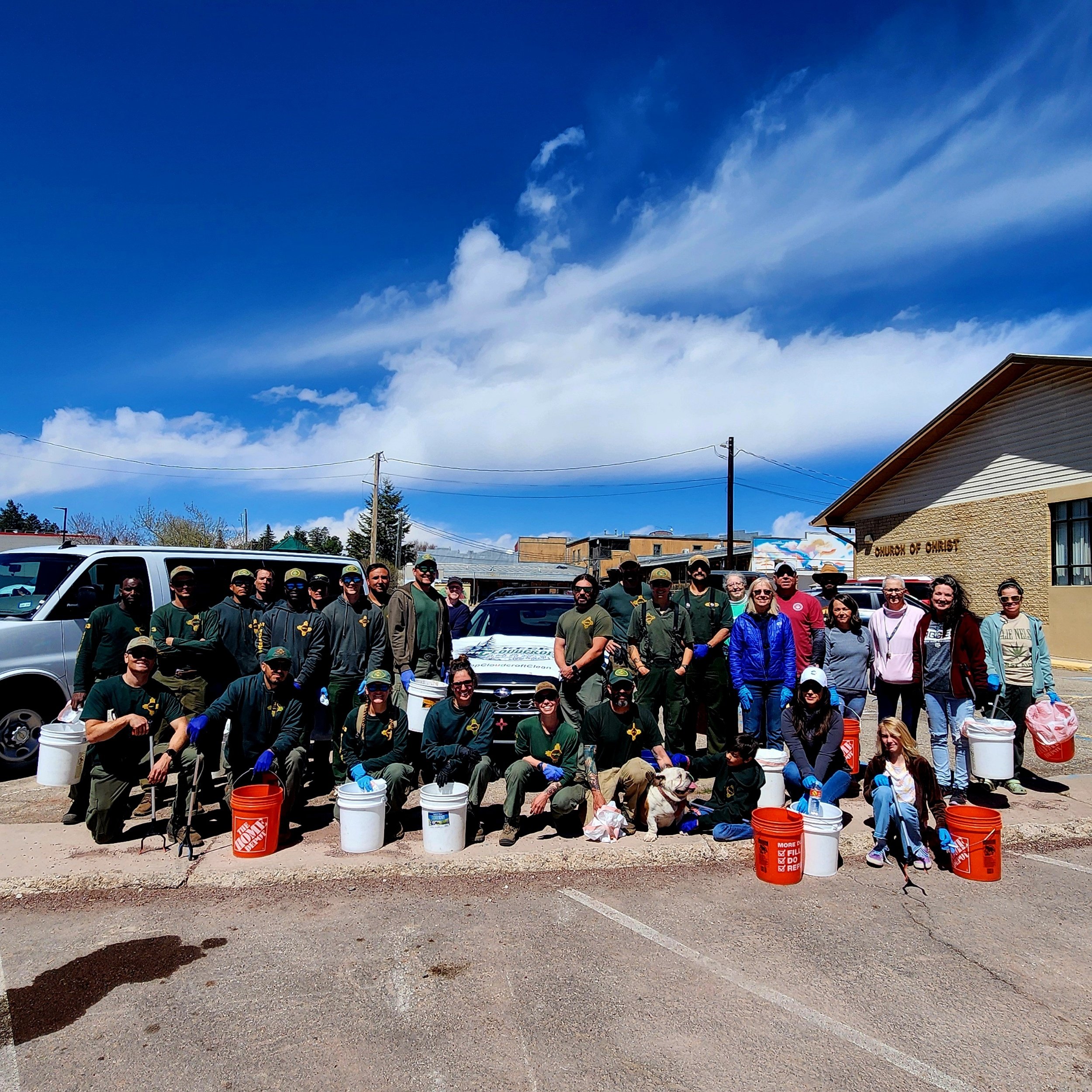 Group of people posing outdoors with cleaning buckets and a dog, in front of vehicles and a building on a sunny day.