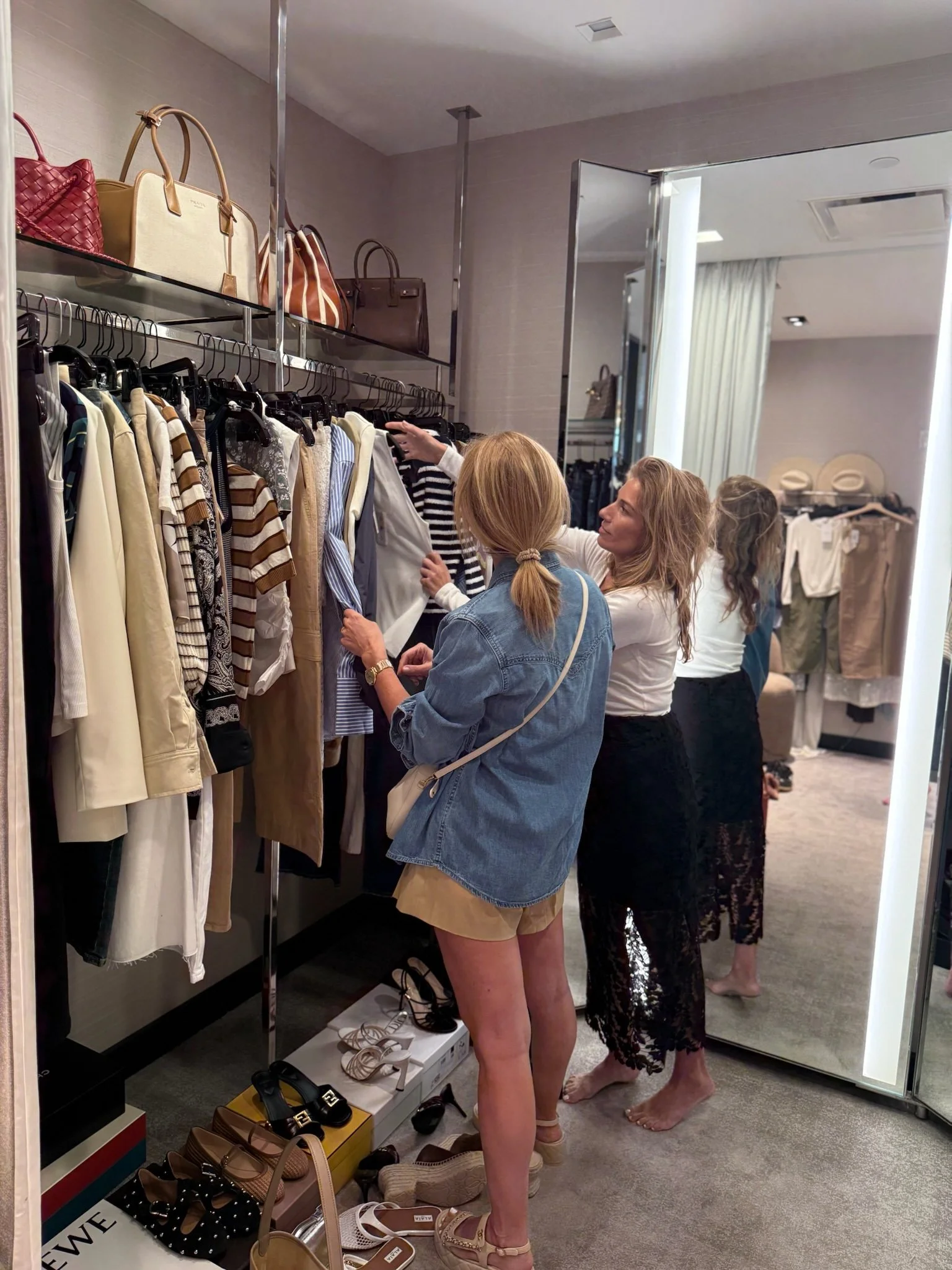 Two women shopping for clothes in a boutique, standing in front of a clothing rack with various garments and handbags displayed on shelves above.