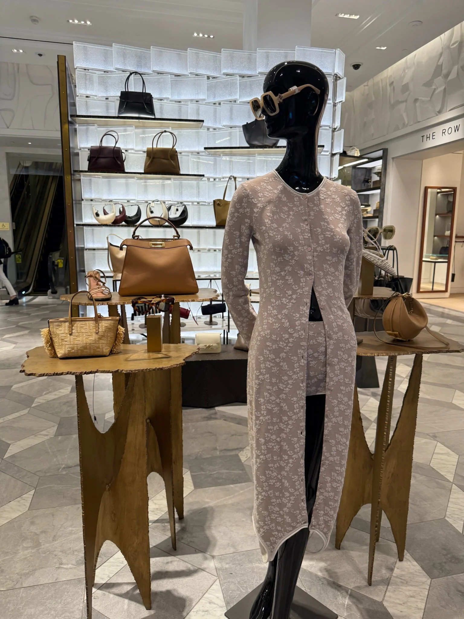 Display of handbags and accessories in a luxury store with a mannequin dressed in a patterned coat and sunglasses.
