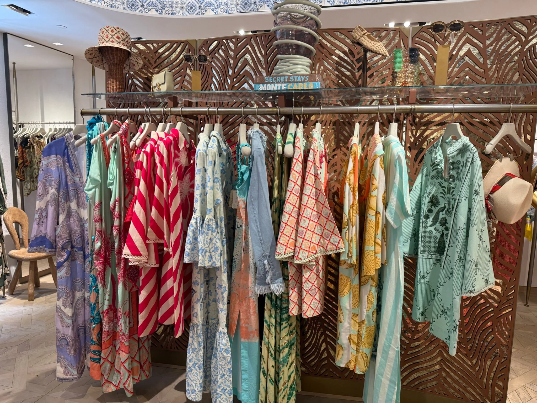 A clothing display in a boutique featuring colorful patterned dresses, shirts, and cover-ups hanging on a rack, with accessories and a decorative wooden panel in the background.
