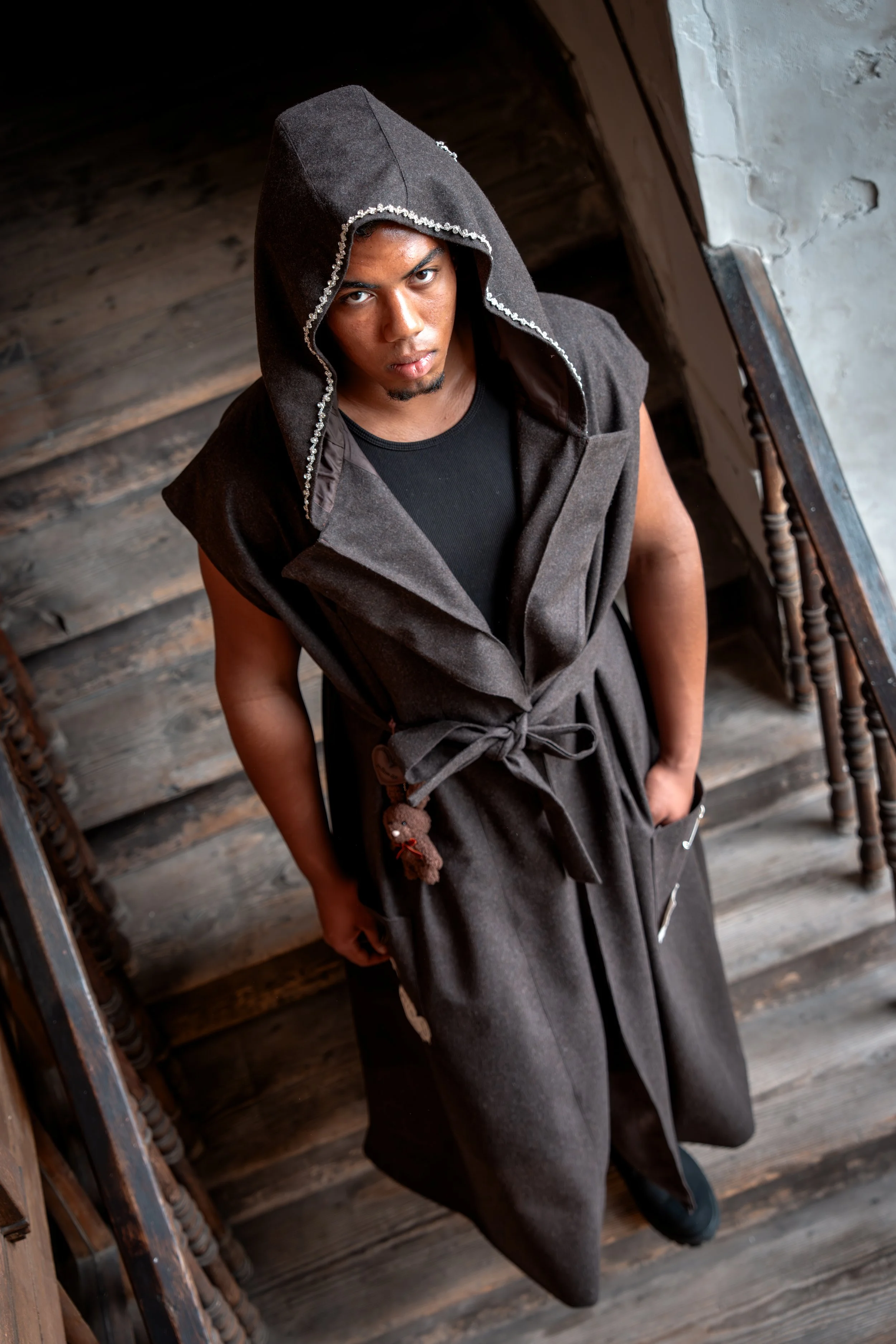 A person wearing a sleeveless black top and a brown hooded vest with a tie waist, standing at the top of a wooden staircase inside a rustic building with wooden floors and walls.