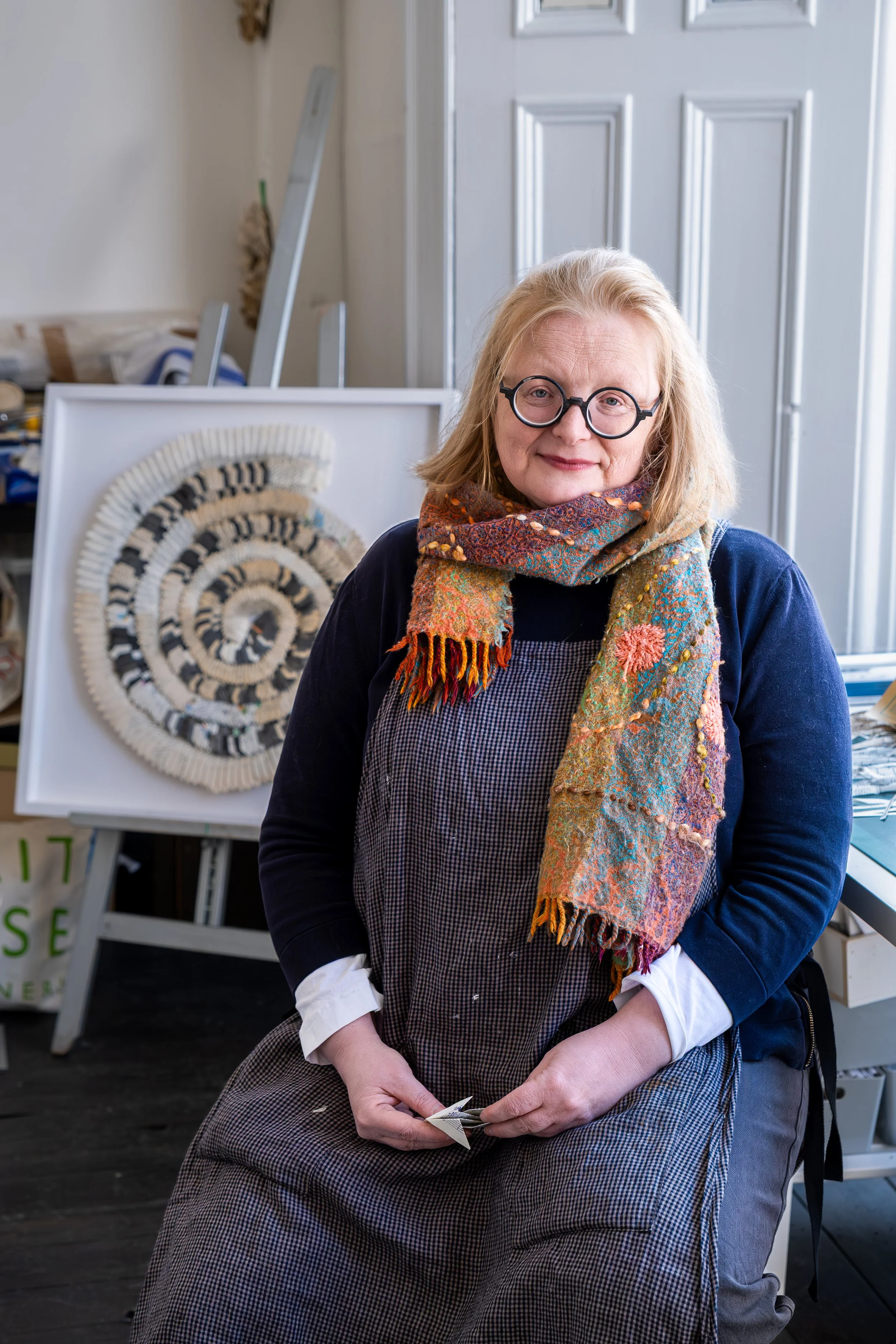 An older woman with blonde hair, glasses, and a colorful scarf sitting on a chair with another person visible in front of her. Behind her, there is a framed piece of textile art with a spiral pattern and some art supplies or artwork in the background.