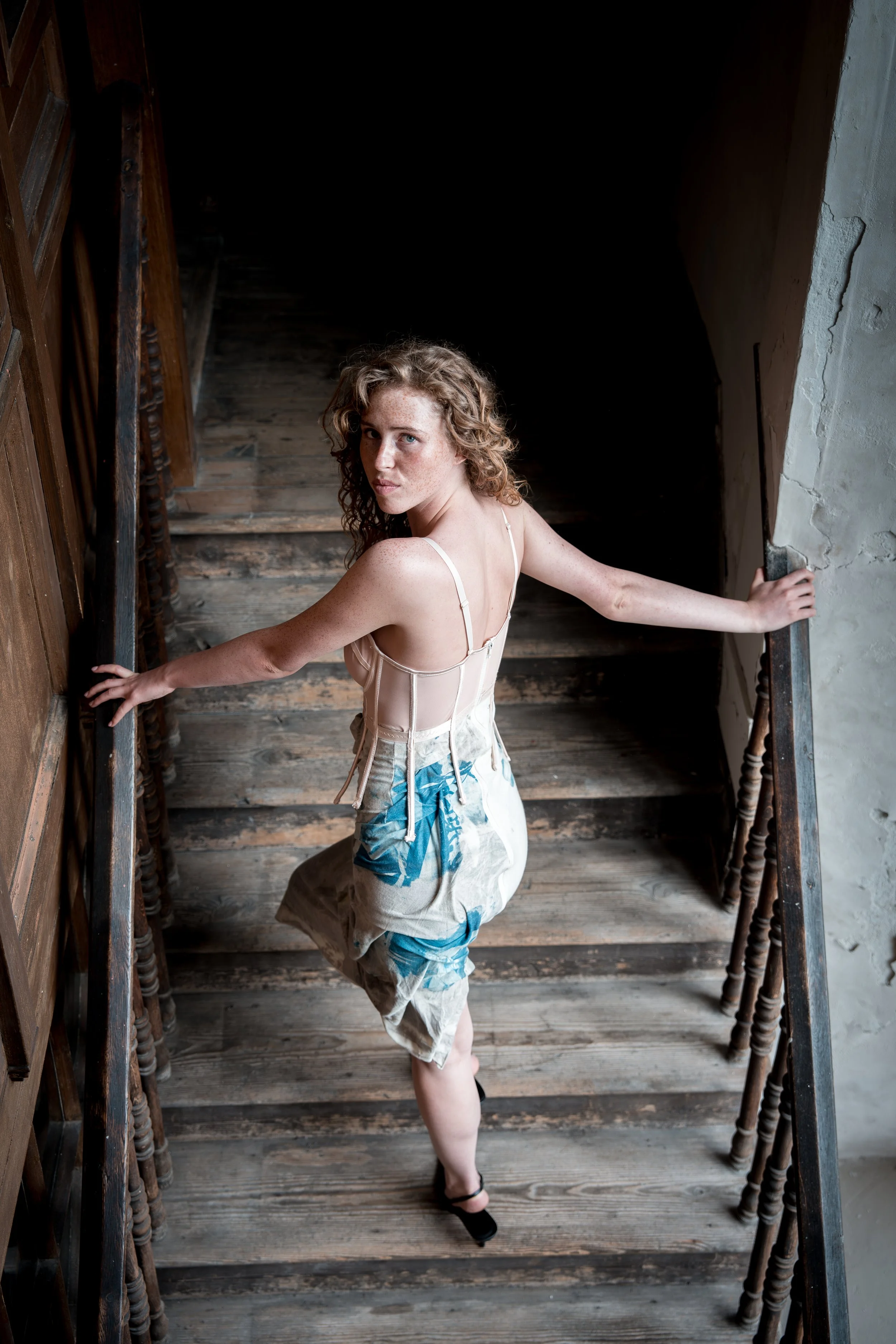 A woman with curly hair and fair skin standing on a wooden staircase, looking up at the camera, wearing a beige corset top, a beige skirt with blue and white floral patterns, and black shoes, holding the staircase railing with both hands.