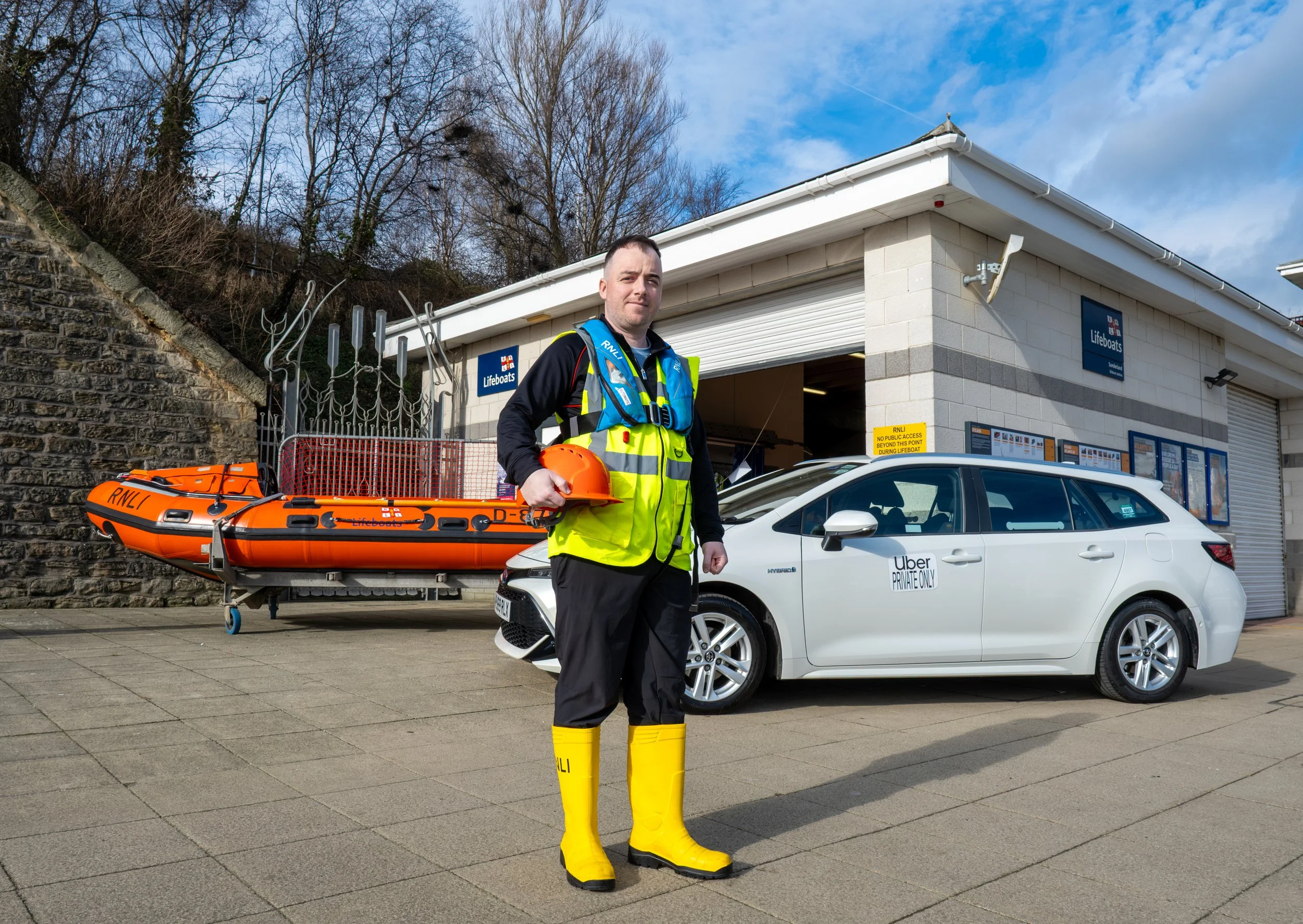 A man in safety gear stands outdoors near a white building with a garage door and signs for lifeboats. He holds an orange helmet and wears bright yellow waterproof boots. An orange lifeboat is secured on a wheeled platform beside him, and a white car
