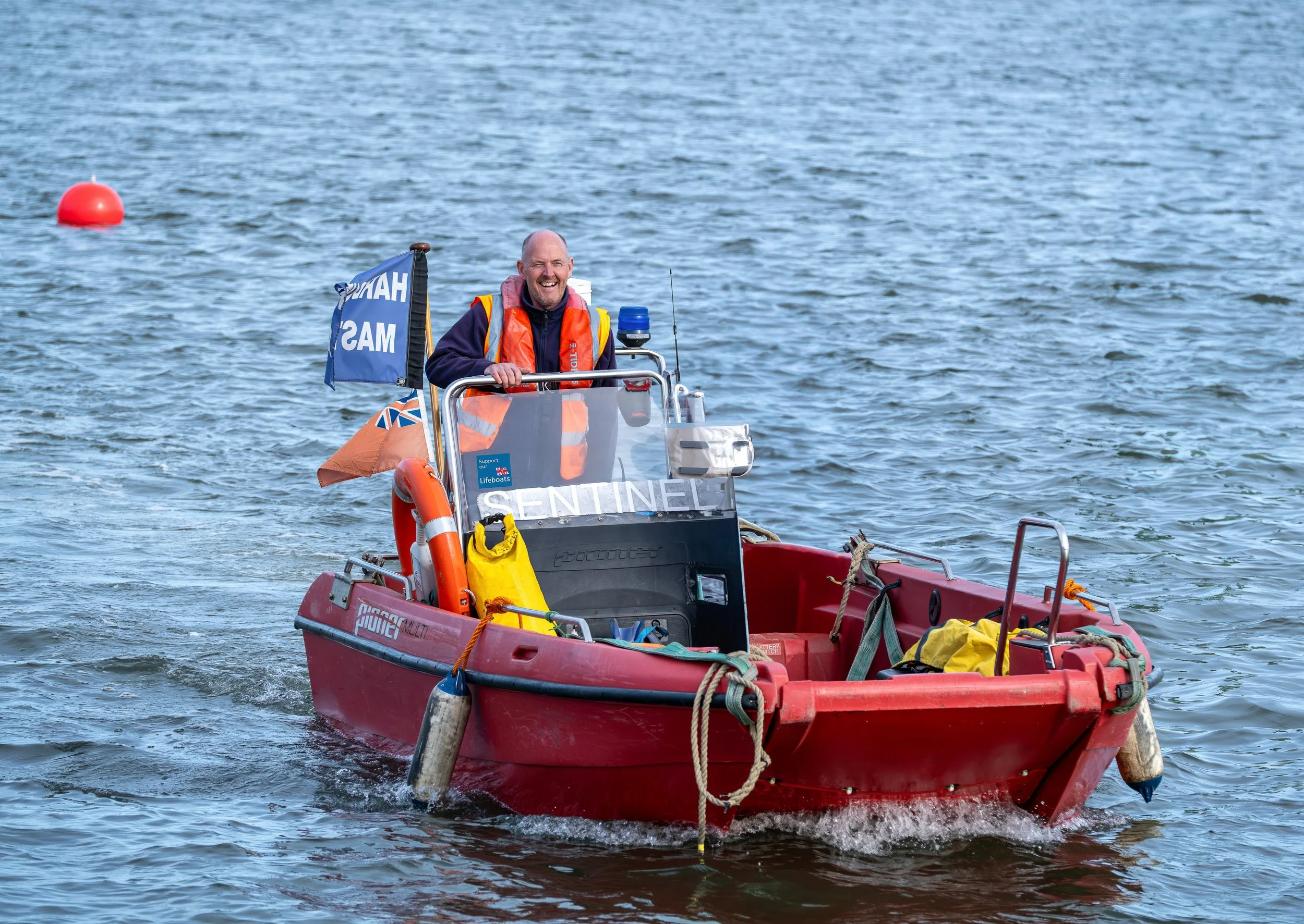 A smiling man in a life jacket rides a red and orange rescue boat on water, with buoys in the background.