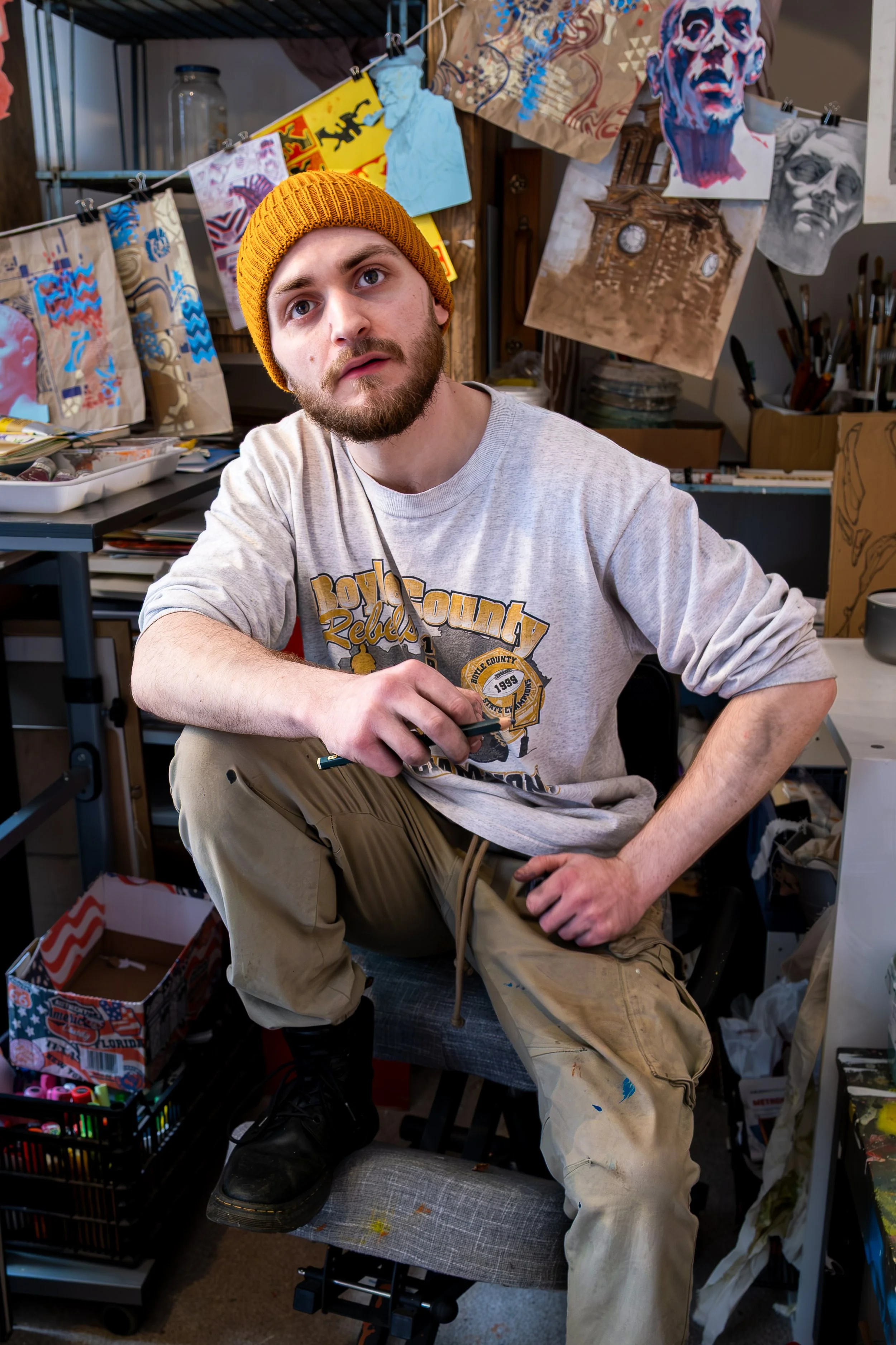 A young man with a beard and a yellow knit cap, sitting in an art studio surrounded by paintings, sketches, and art supplies, holding a paintbrush.