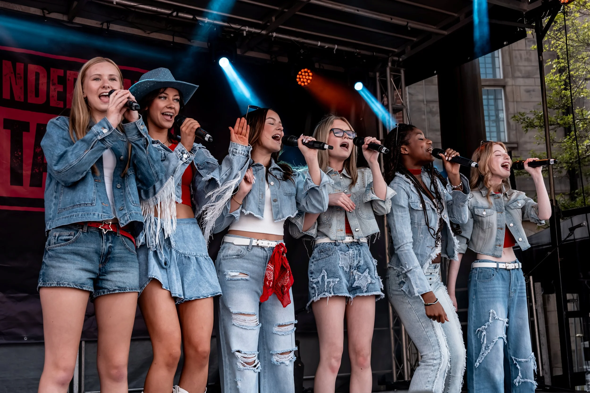 Six young women are performing on an outdoor stage, singing into microphones and wearing denim jackets and ripped jeans.