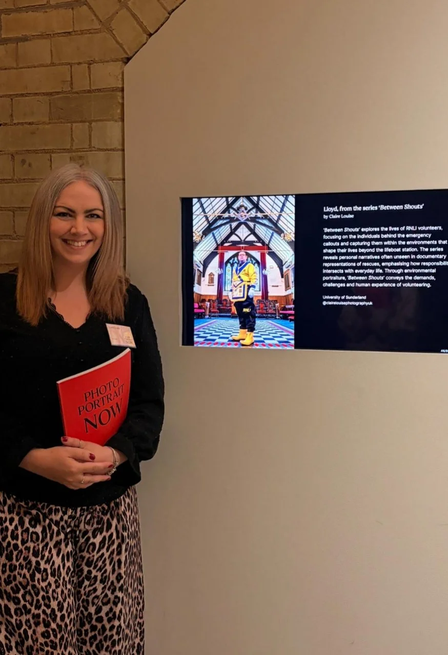 Woman standing next to a display screen, holding a red booklet titled "Photo Portrait Now". The display screen shows information about a documentary series called "Between Shouts" with an image of a performer in a church-like setting.