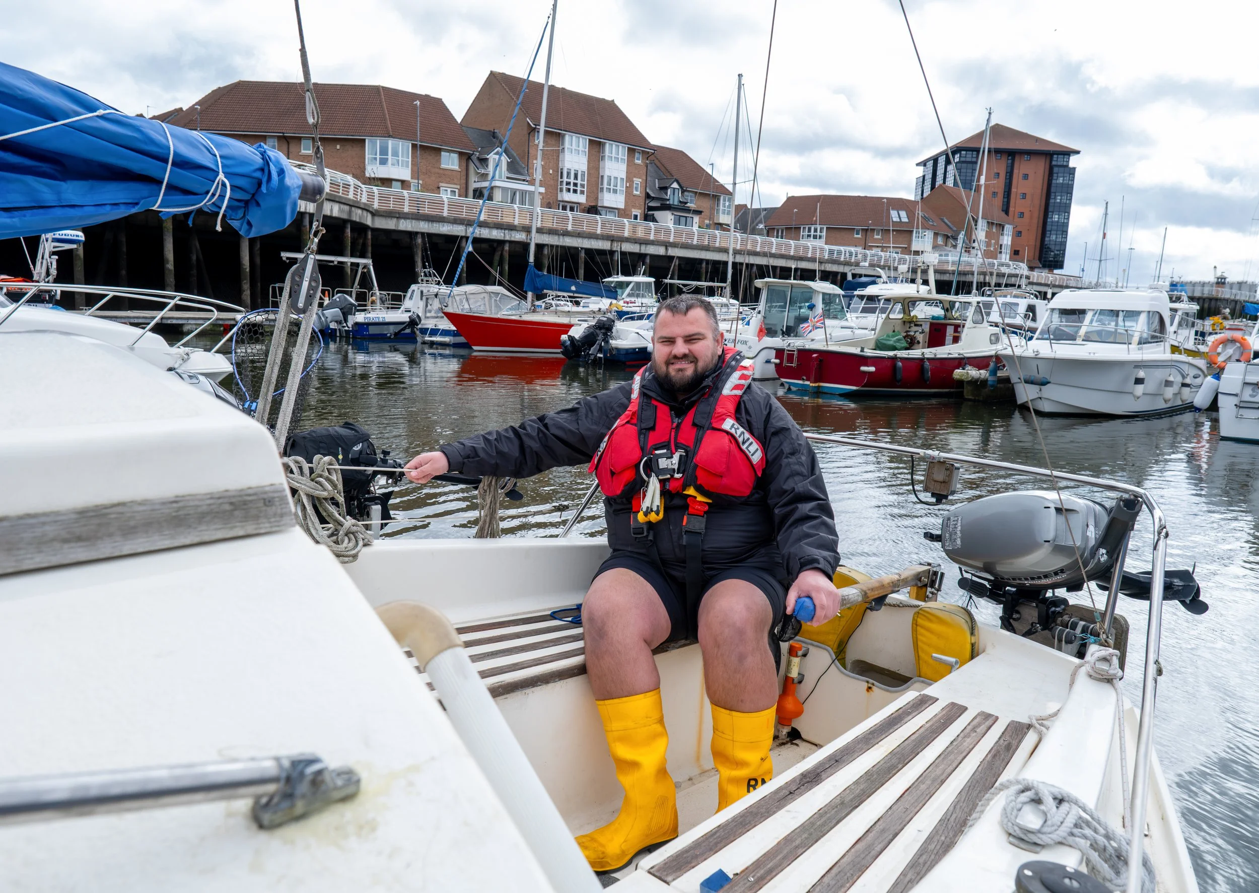 A man sitting on a small boat in a marina, wearing a black jacket, red life vest, yellow waterproof boots, and smiling.