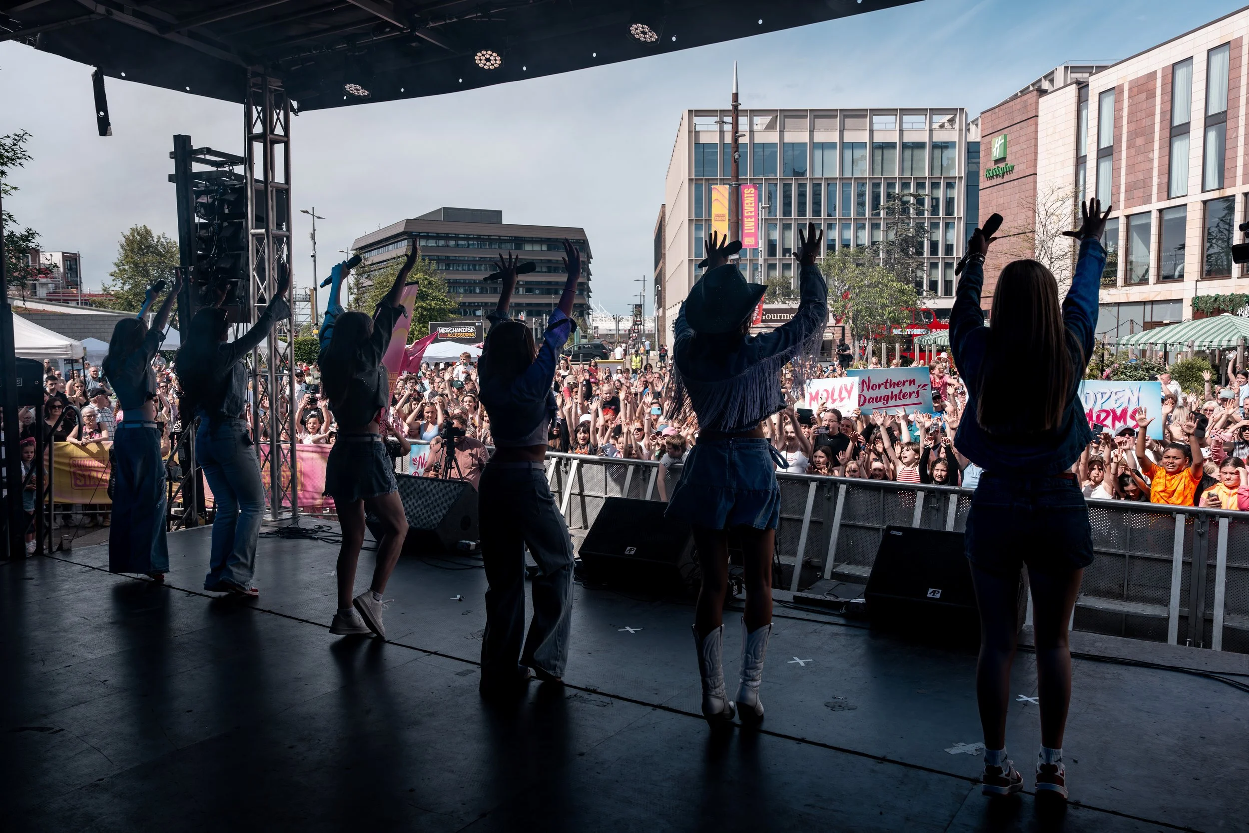 Performers on stage entertaining a large outdoor crowd during a daytime event in an urban area.