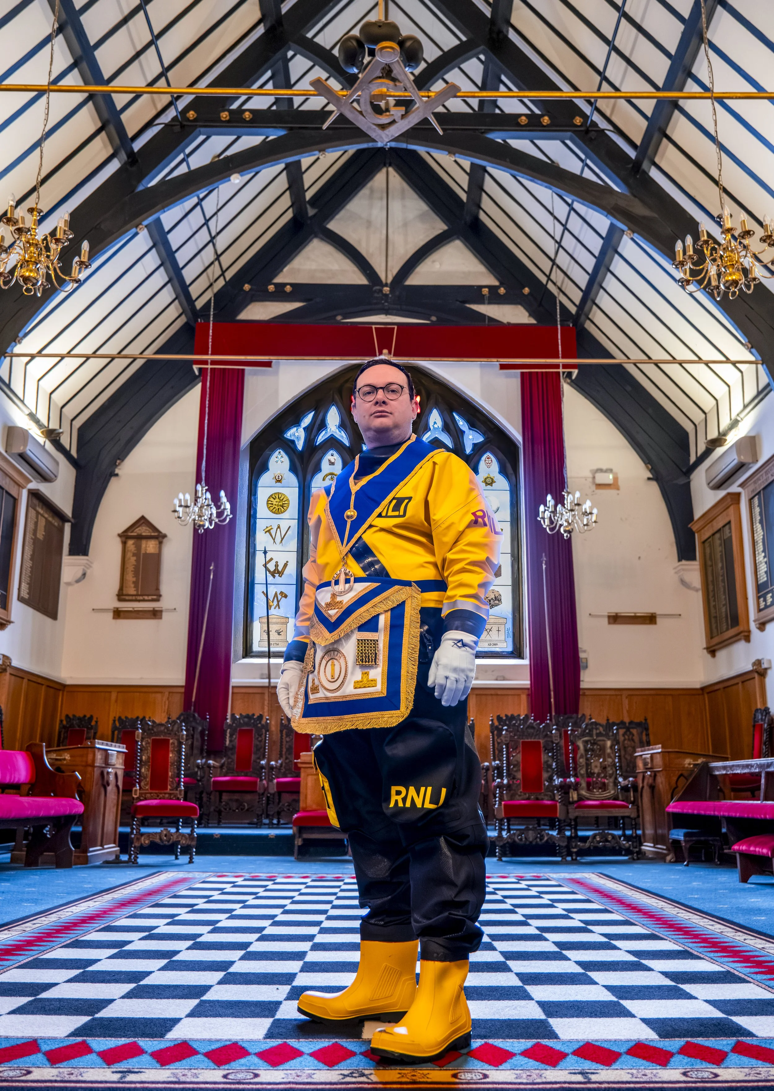 A person dressed in U.S. national language (RNL) uniform, including a yellow and black jacket, yellow rubber boots, and white gloves, is standing inside a church or cathedral with stained glass windows and red curtains. The person faces the camera wi