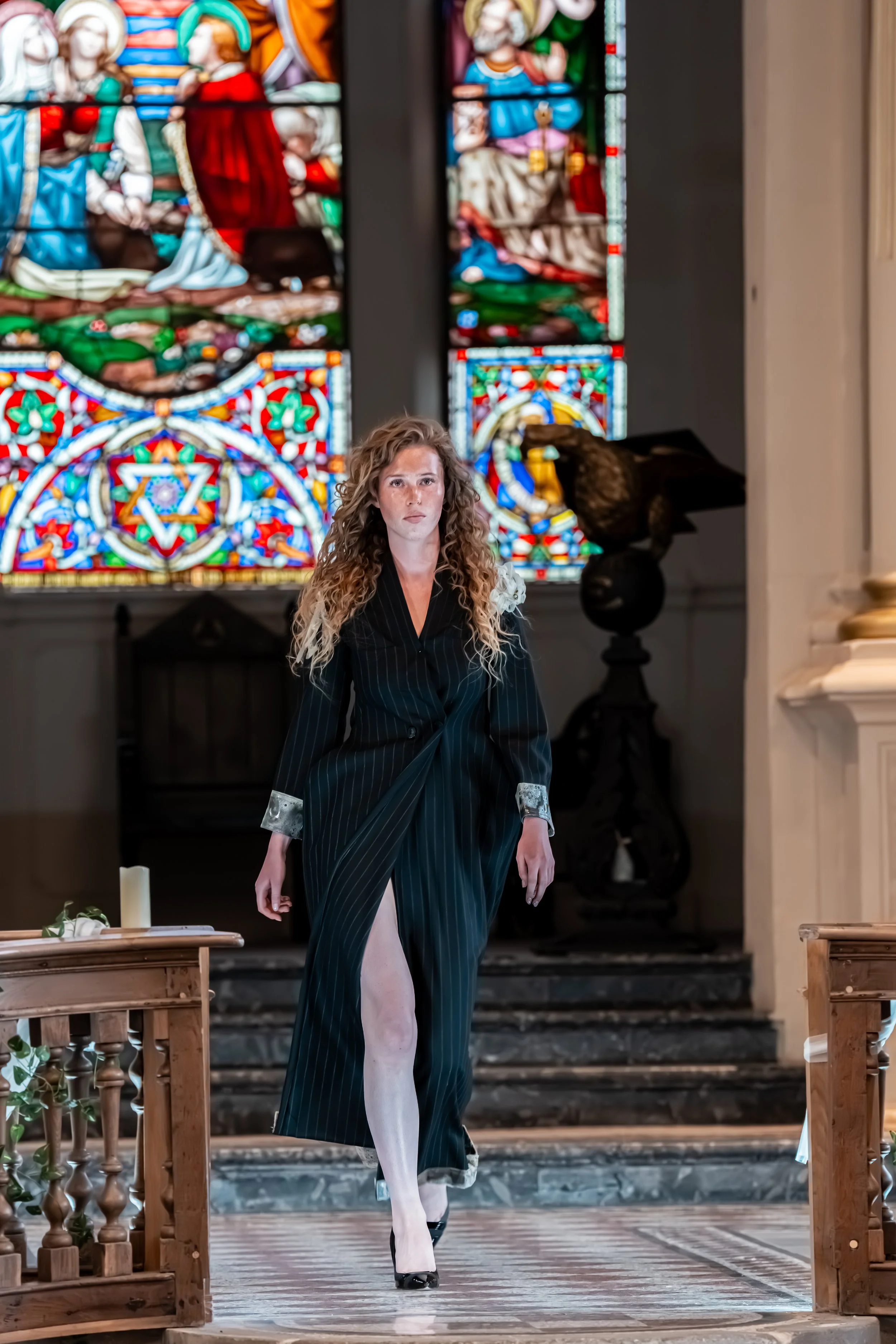 A woman with curly hair walking down the aisle of a church, wearing a long, dark-striped dress with an open slit and high heels, in front of colorful stained glass windows.