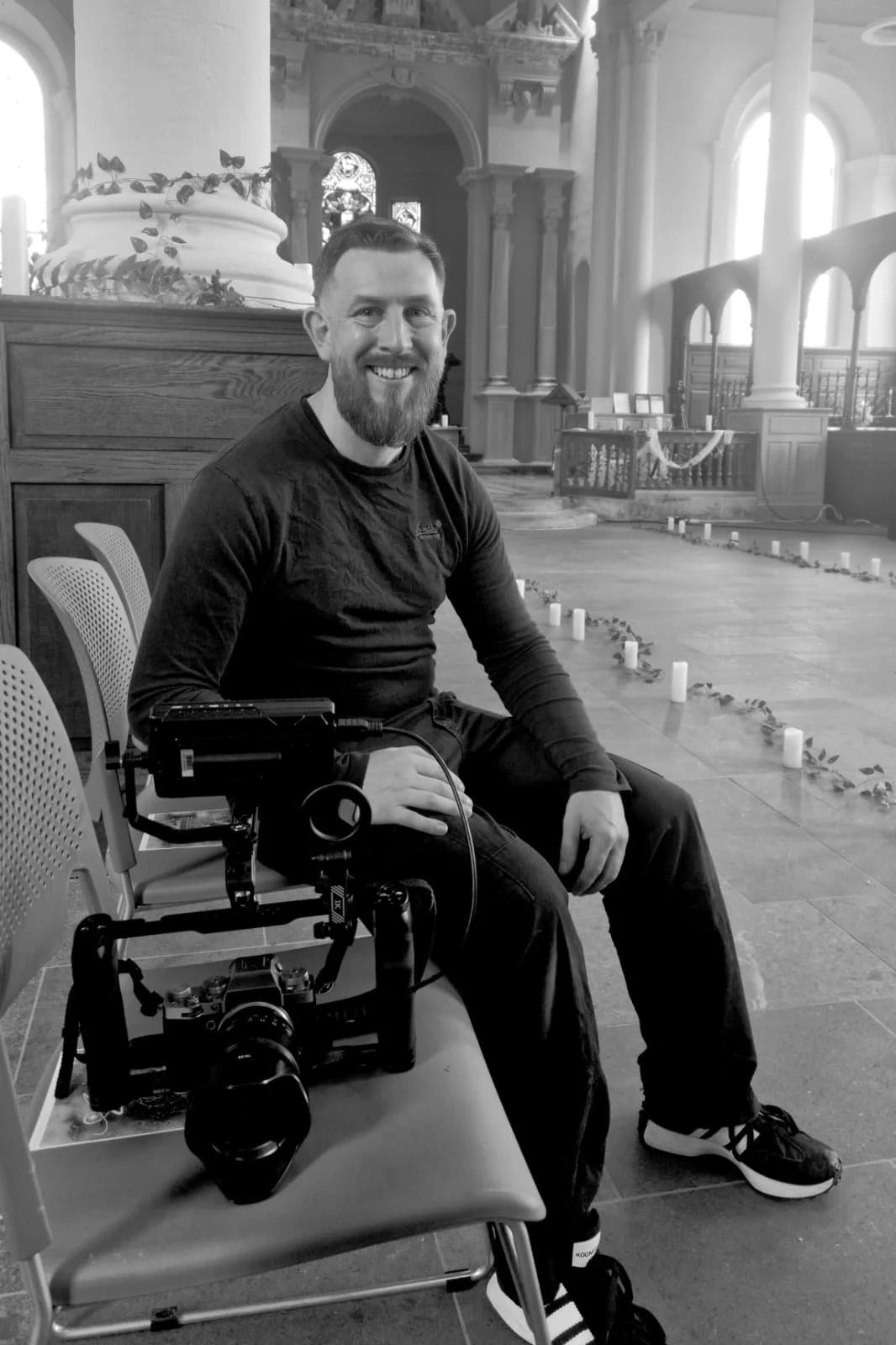 A smiling man in black clothing sitting in a church, with candles and flower decorations on the floor behind him.