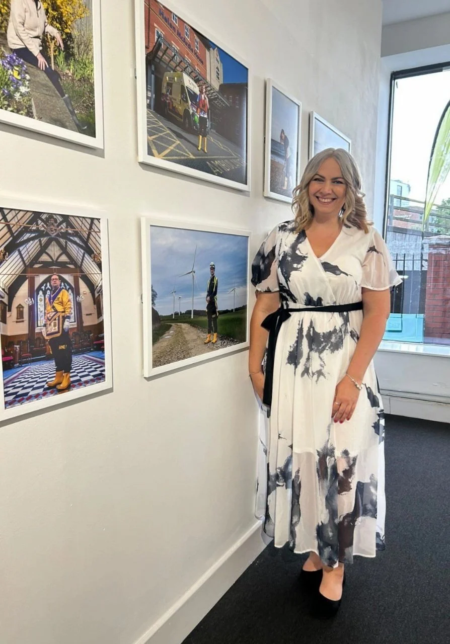 A woman smiling in a white and black floral dress standing in front of a wall with framed photographs.