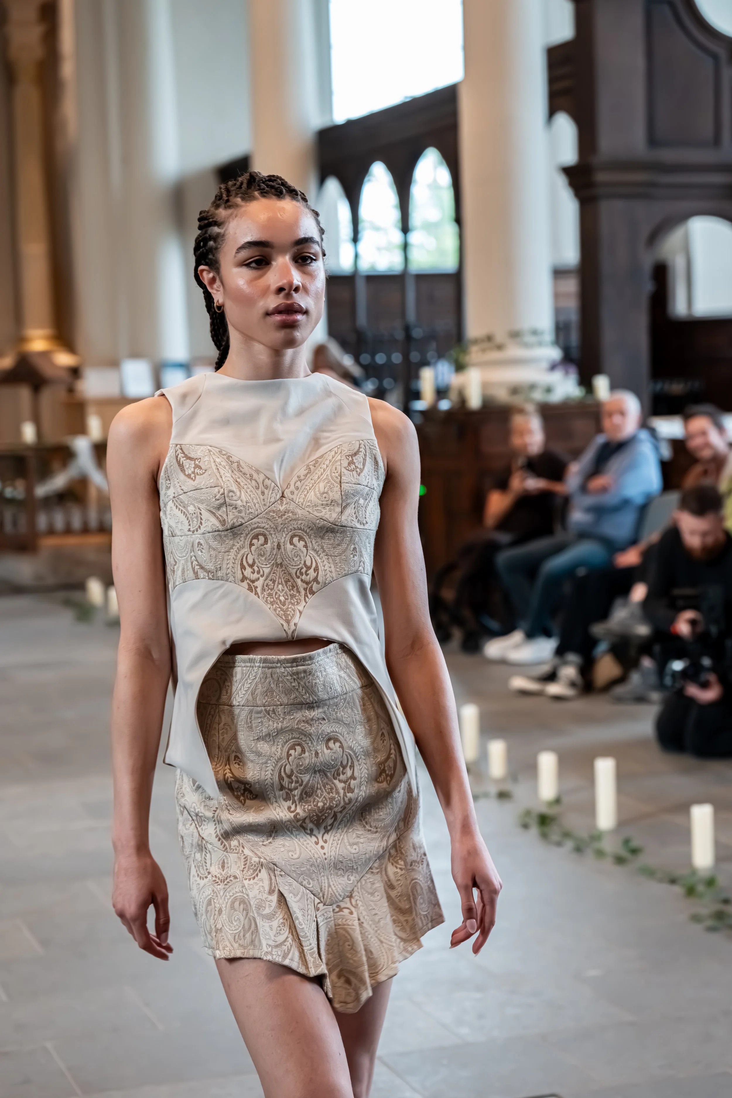 A young woman walking in a church wearing a beige, intricate-patterned dress. There are candles and people seated in the background, with large windows and wood accents visible.