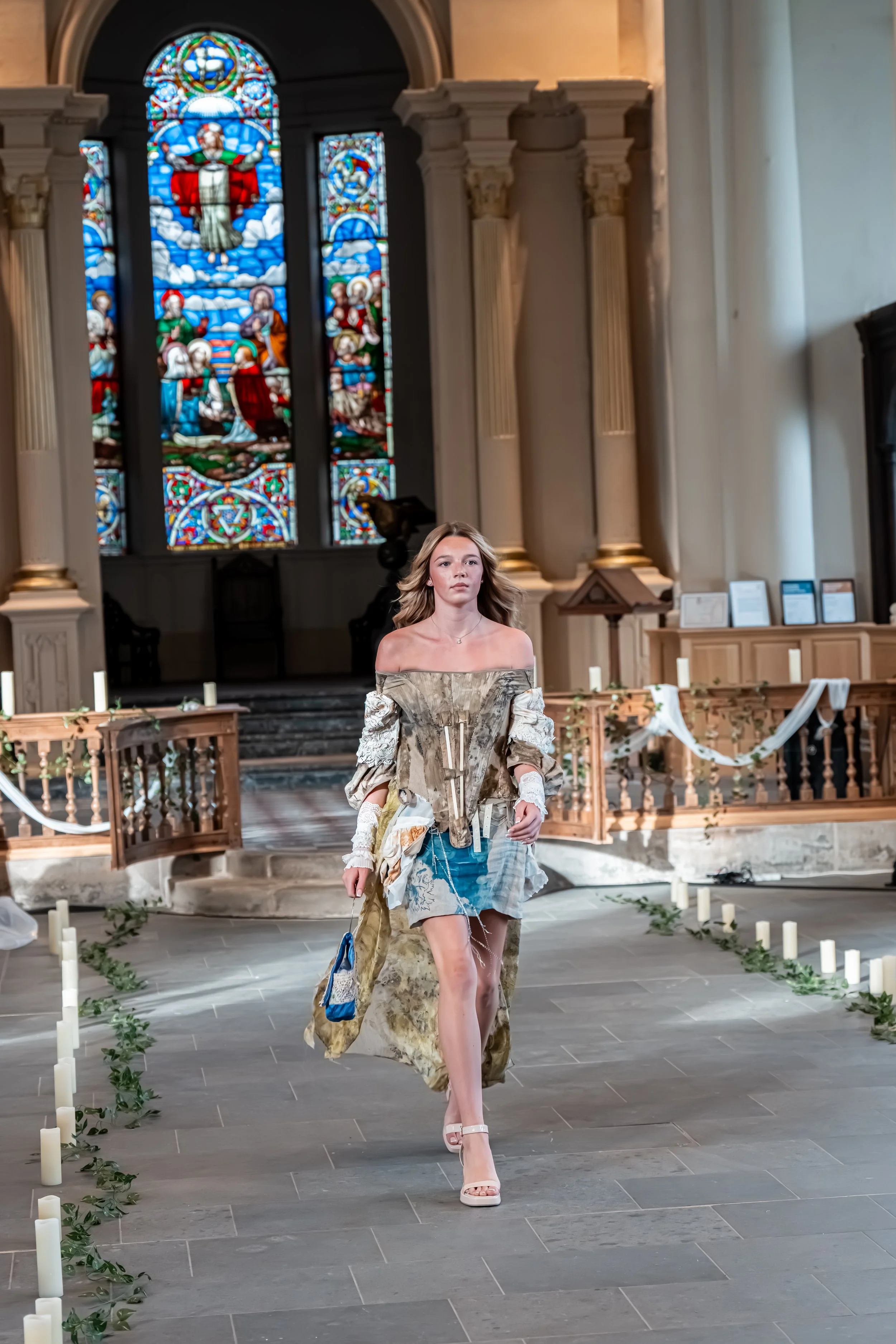 A woman walking on a cathedral aisle with stained glass windows in the background, candles and greenery along the floor.