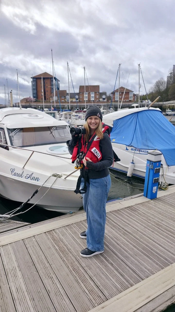 A woman standing on a marina dock, holding a camera, smiling, wearing a beanie, dark hoodie, jeans, and sneakers, with boats and buildings in the background.