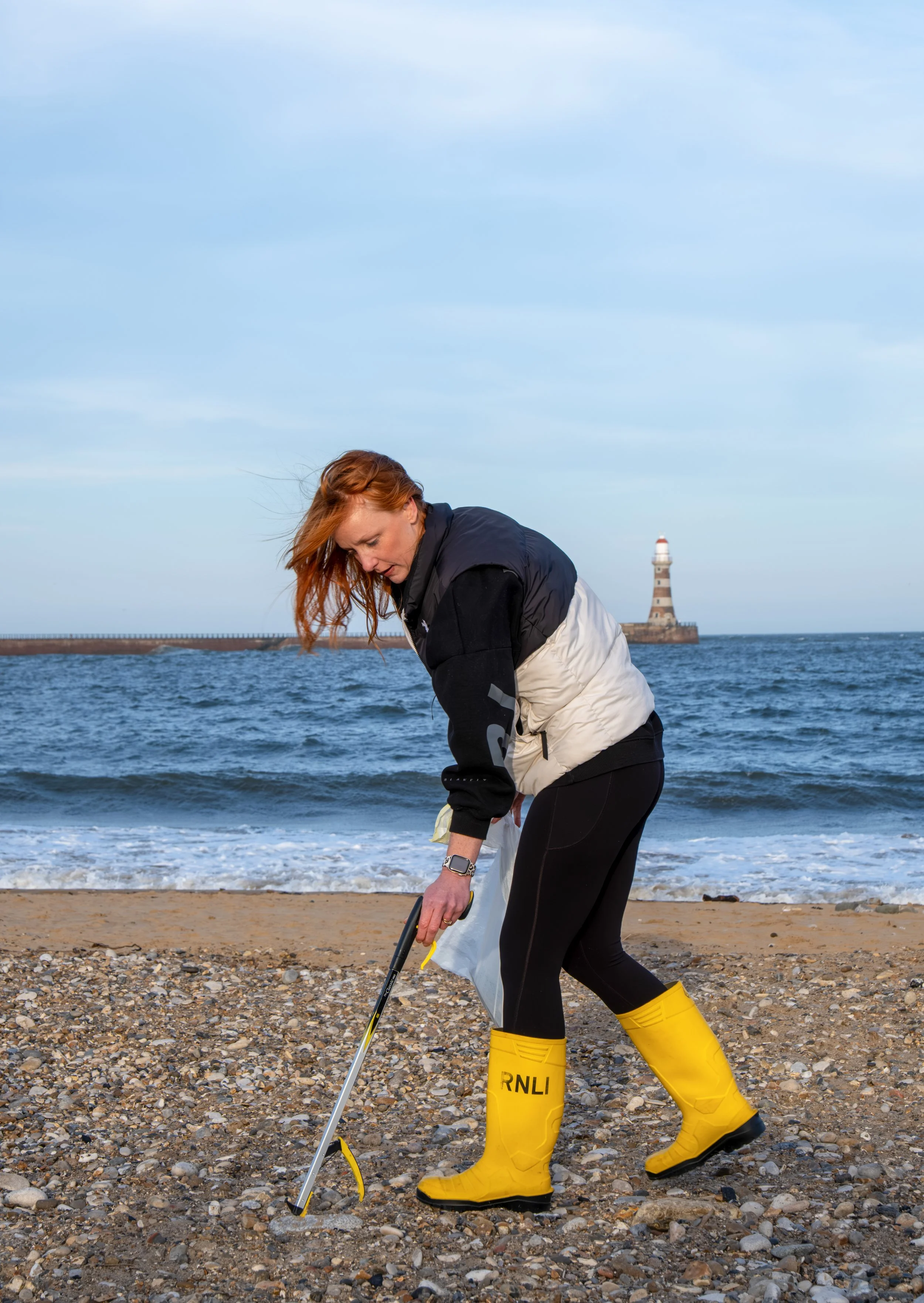 A woman in yellow waterproof boots labeled 'RNLI' is picking up trash on a pebbled beach near the ocean with a lighthouse in the background.