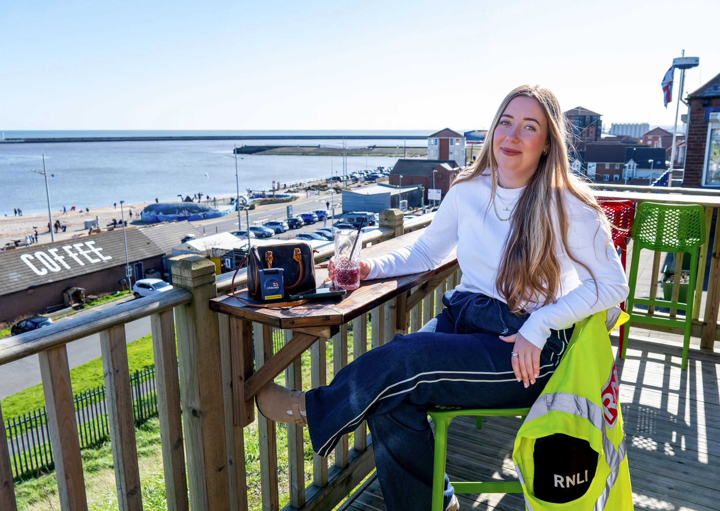 A woman with long hair sitting on a green chair on a balcony overlooking a beach, holding a drink, with a coast and cars in the background during daytime.