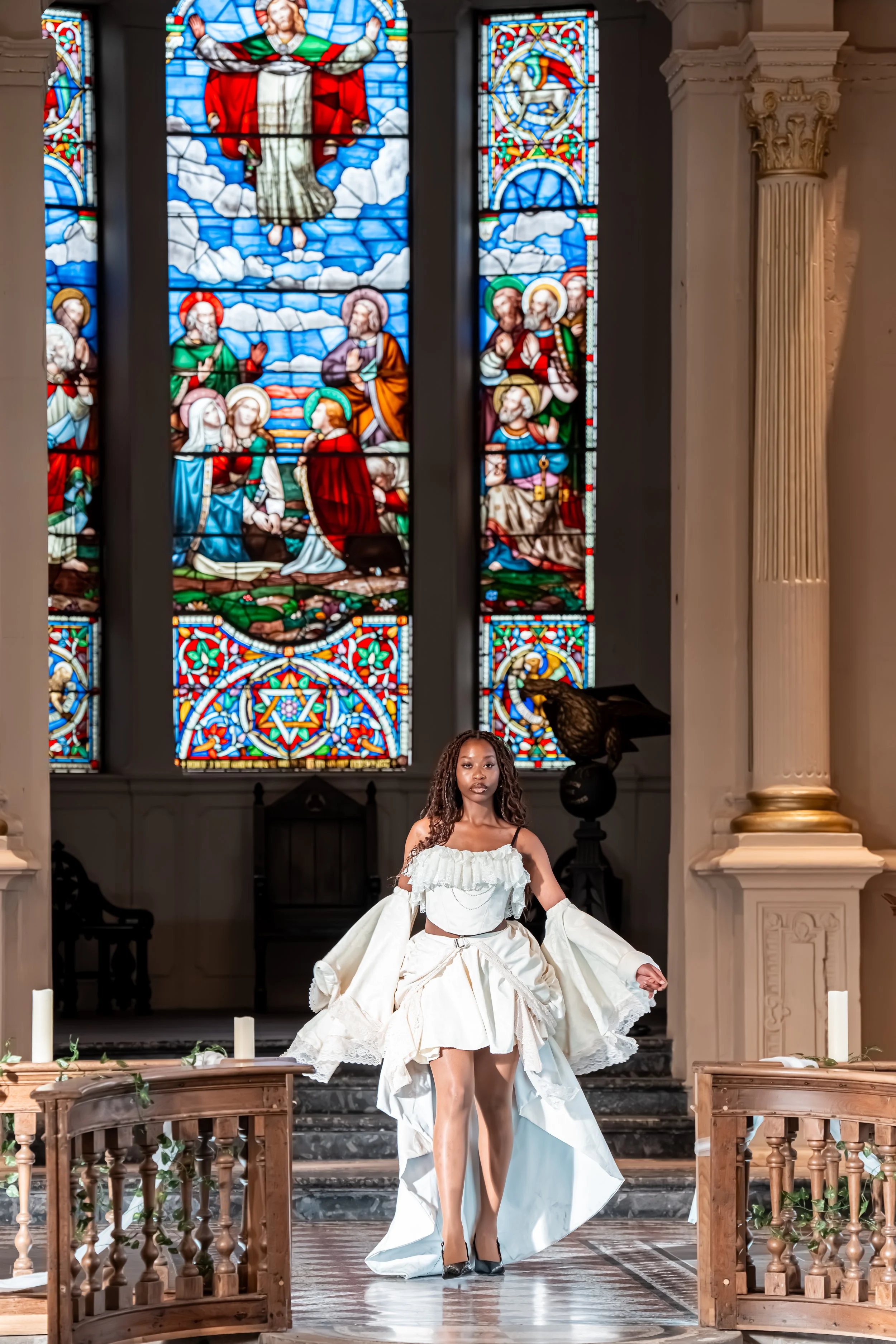 A woman in a white dress standing in front of a stained glass window inside a church. The window depicts a religious scene, and there are candles and a wooden railing in the foreground.