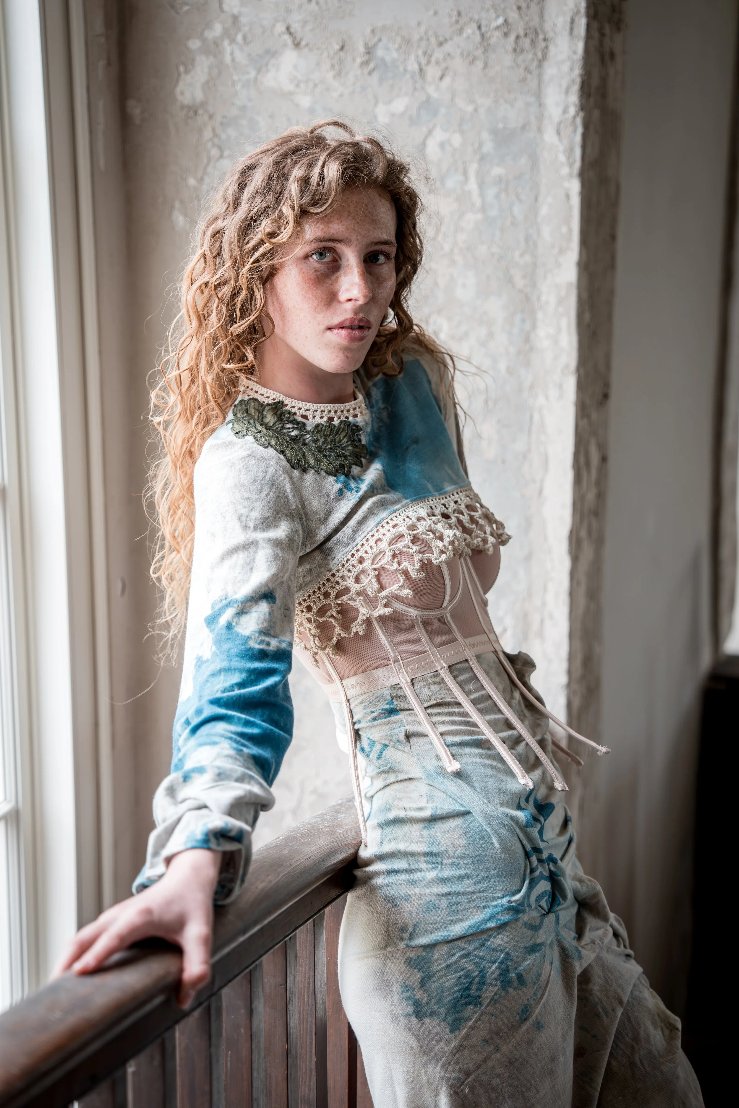 A woman with curly red hair and freckles sitting on a wooden railing by a window in a rustic setting, wearing a patchwork blouse and distressed jeans.