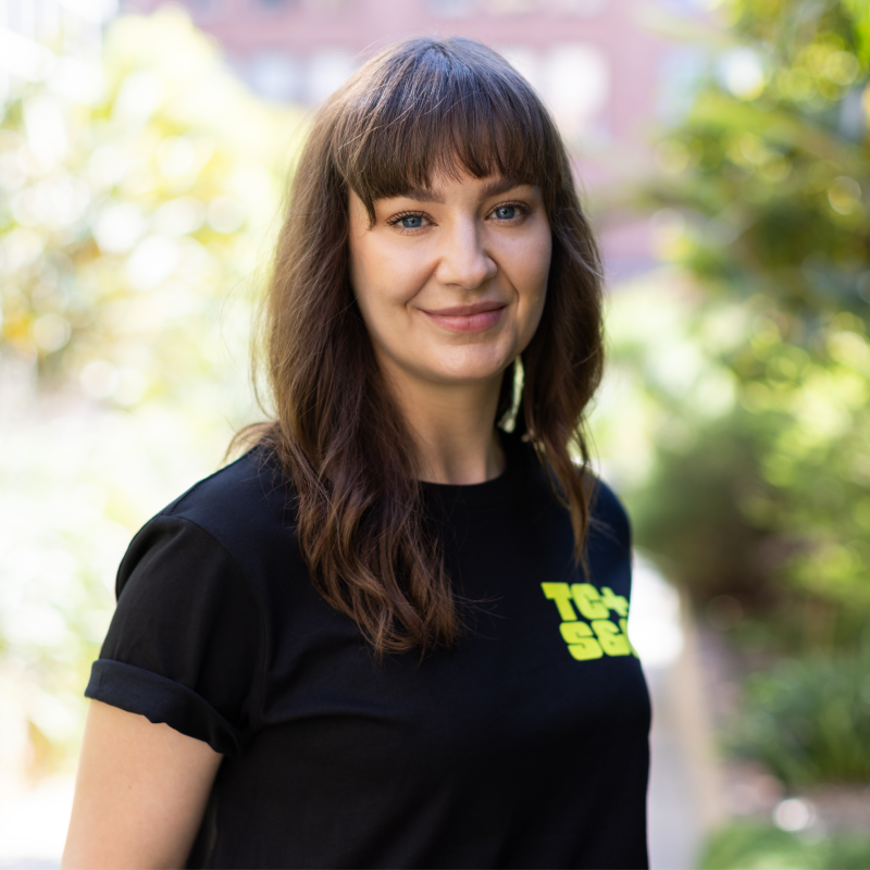 Young woman outdoors with brown wavy hair wearing a black T-shirt with yellow text, smiling at the camera, with blurred greenery in the background.