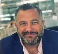 A man with short gray hair and a beard, wearing a dark blazer and a light-colored shirt, smiling at the camera.