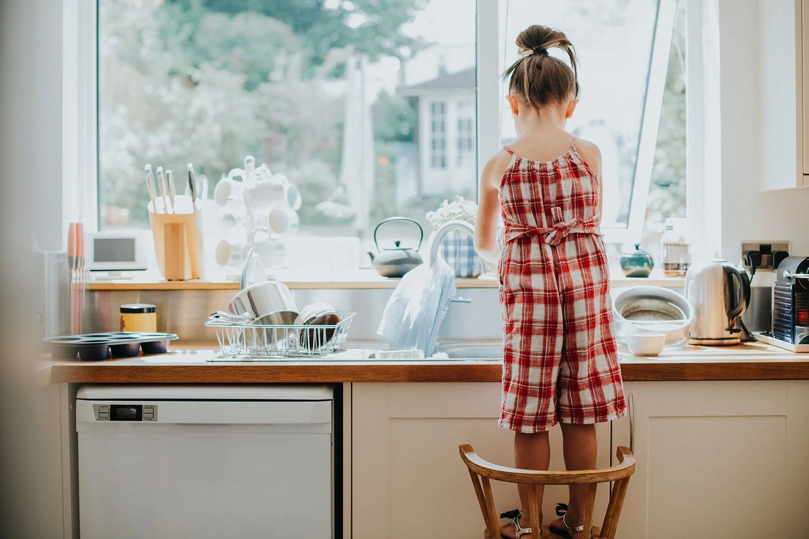 Little girl doing dishes independently while standing on stool at kitchen sink.