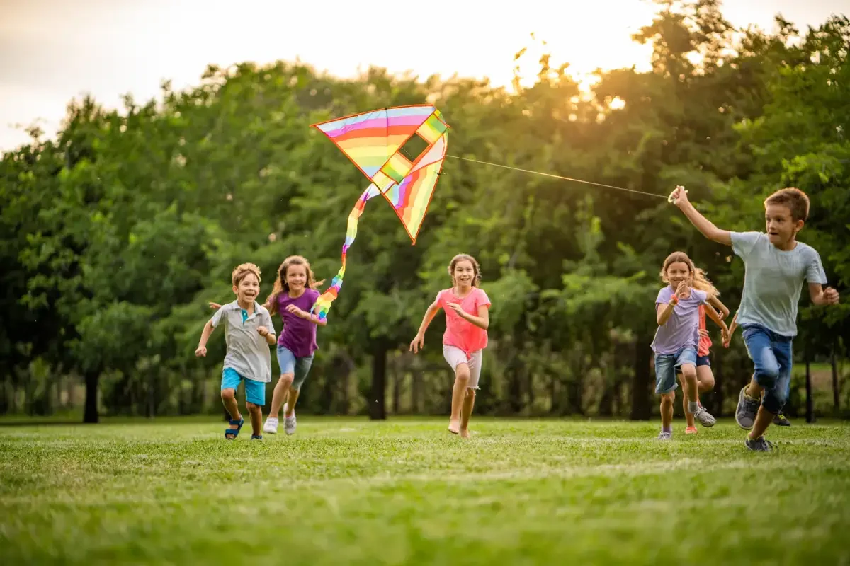 A group of five children laughing and running across a green grassy field while flying a colorful rainbow kite at sunset.