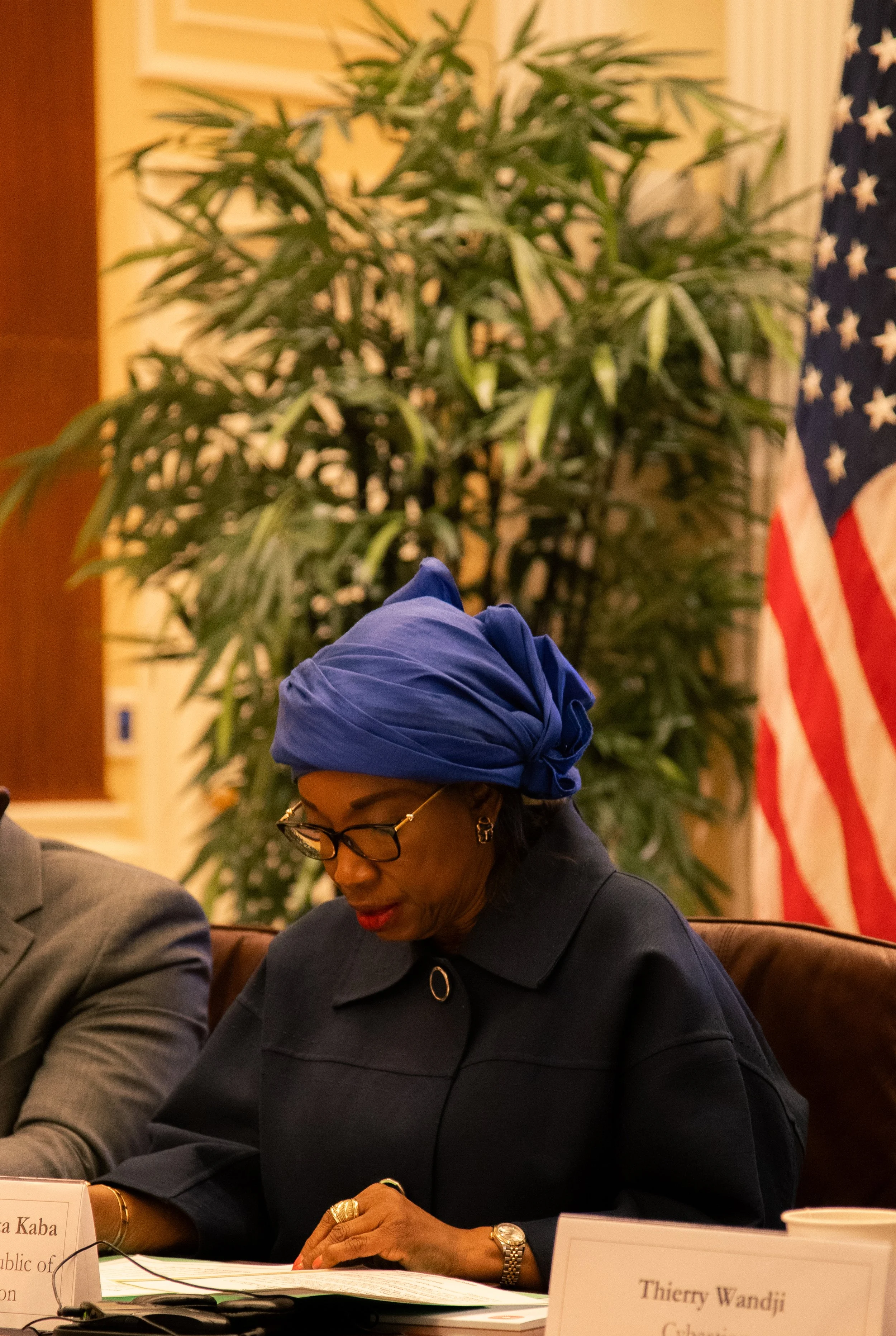A woman wearing a blue headwrap, glasses, and a dark blazer sitting at a conference table with a nameplate that reads 'Thierry Wandji' and the text 'Republic of' visible. She is looking down at a document on the table. An American flag and green plan
