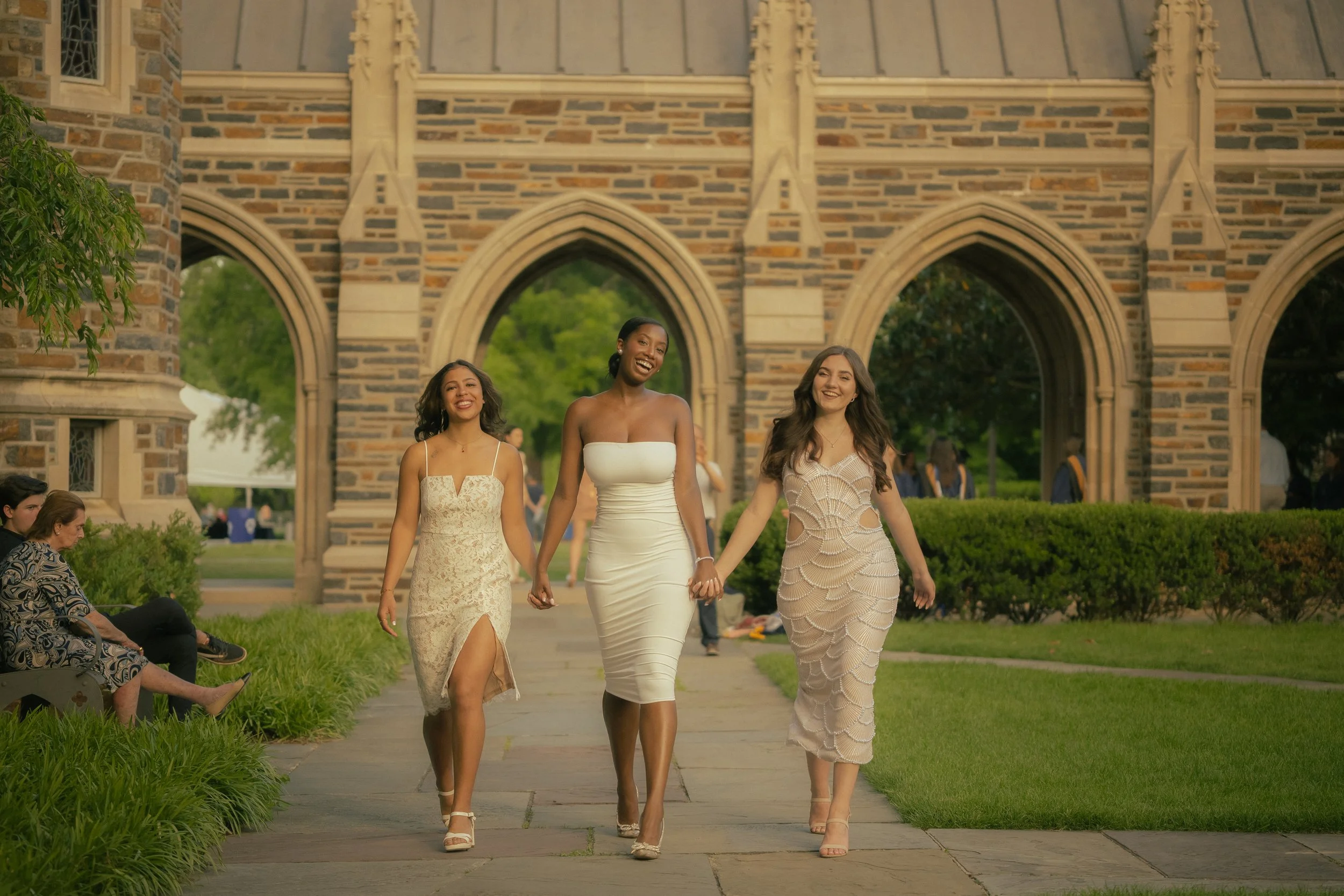 Three women in elegant dresses are walking hand in hand on a sidewalk in front of a historic stone building with arched windows. They are smiling and appear happy. There are people sitting on the grass and trees in the background.