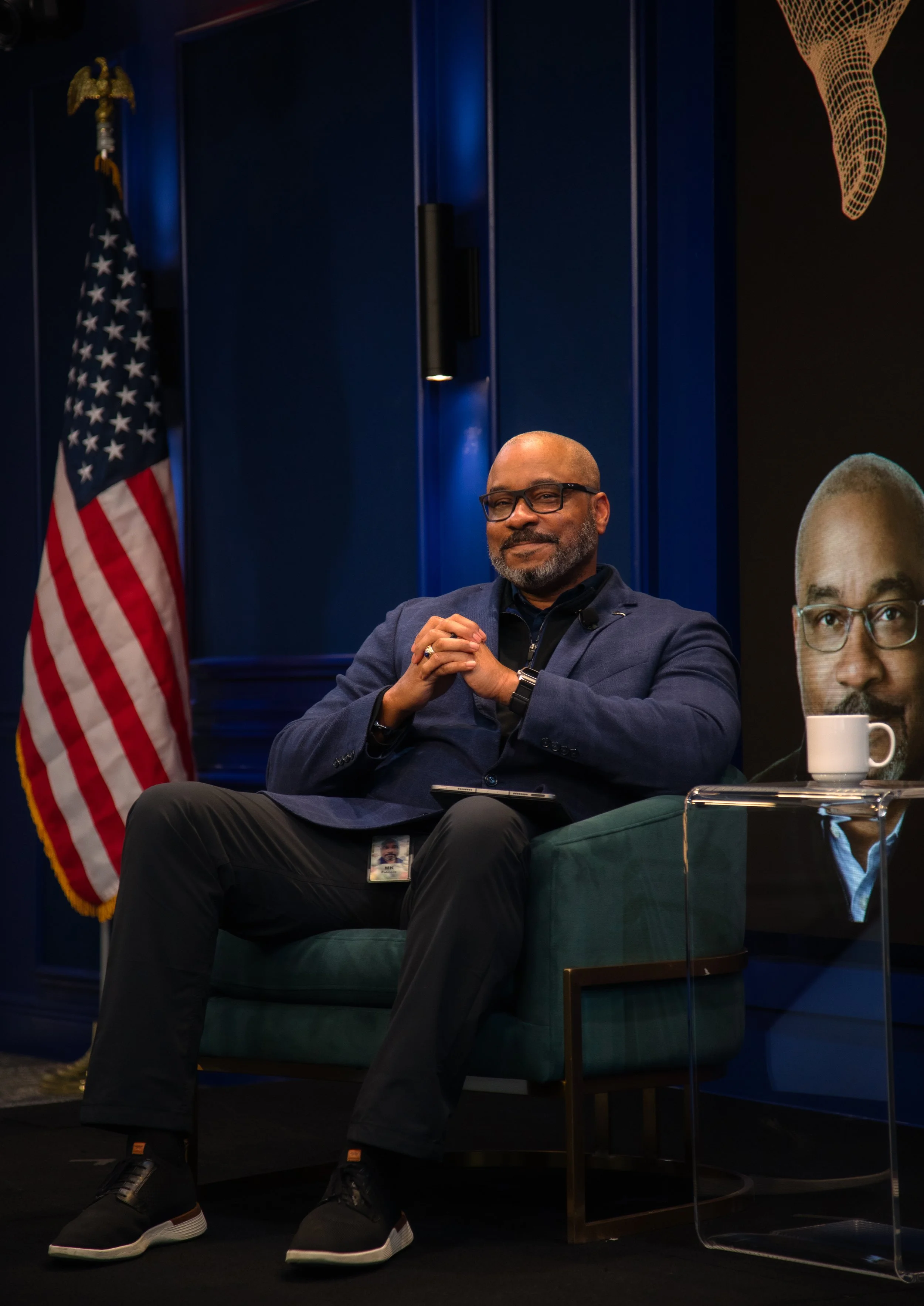 A man with glasses and a beard sitting on a green chair, wearing a navy blazer, black shirt, and black sneakers, with an American flag in the background and a large screen displaying a photo of a man with glasses and a mustache, a white mug on a smal