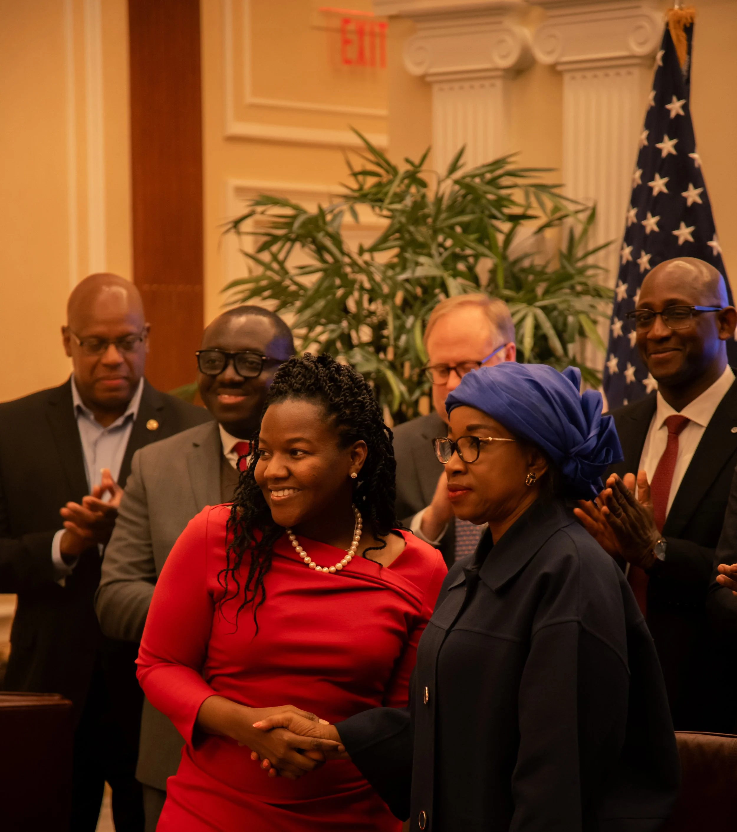 A group of people, including men and women of diverse backgrounds, are standing closely together in a formal setting. One woman in a red dress and pearl necklace is holding hands with a man in a black suit, who is also holding her hand. Other individ