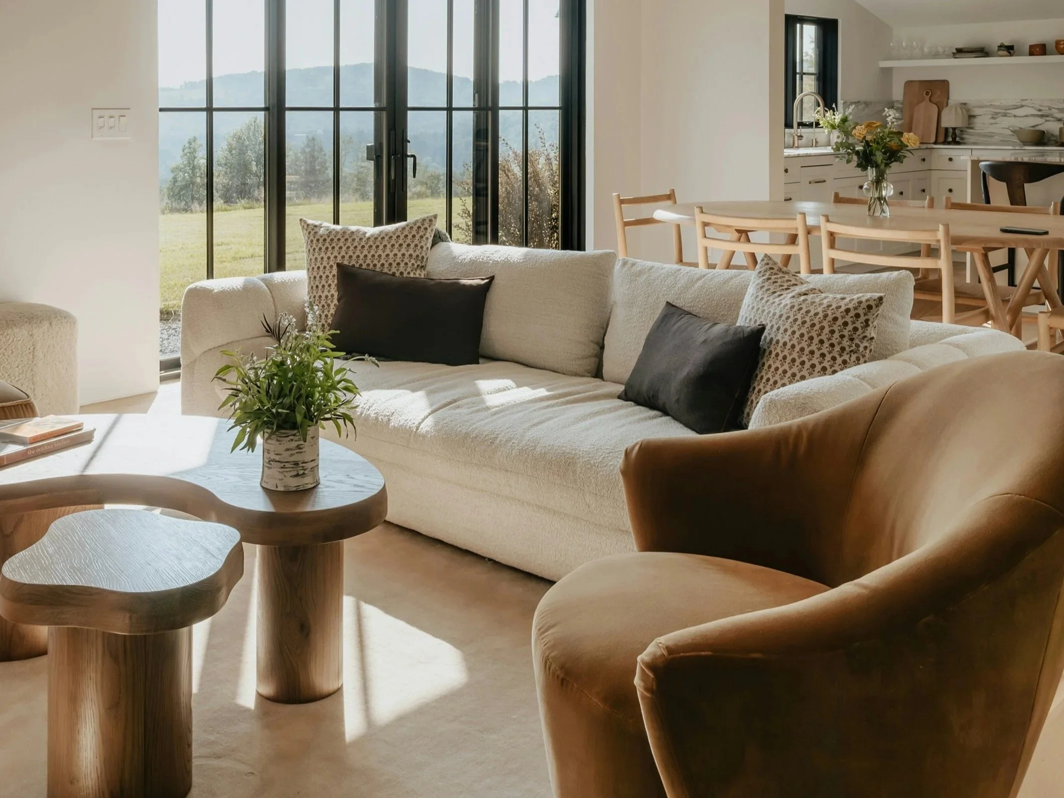 Living room with a cream sofa, black and patterned throw pillows, wooden side tables, and an armchair. Large glass sliding doors reveal a scenic view of a green field and hills outside.