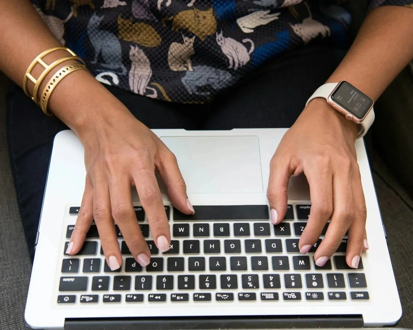 Person typing on a laptop keyboard, wearing a smartwatch on their right wrist and gold bracelets on their left wrist, dressed in a shirt with cat and mouse patterns.