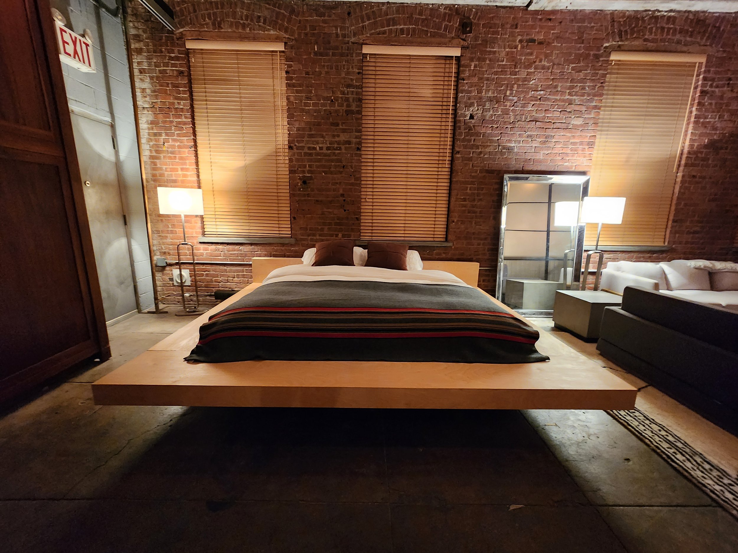A bedroom with a modern, minimalist design featuring a large wooden bed with a low profile, a white couch on the right, and an industrial brick wall background. There are two tall windows with wooden blinds, and two tall lamps with white shades flank