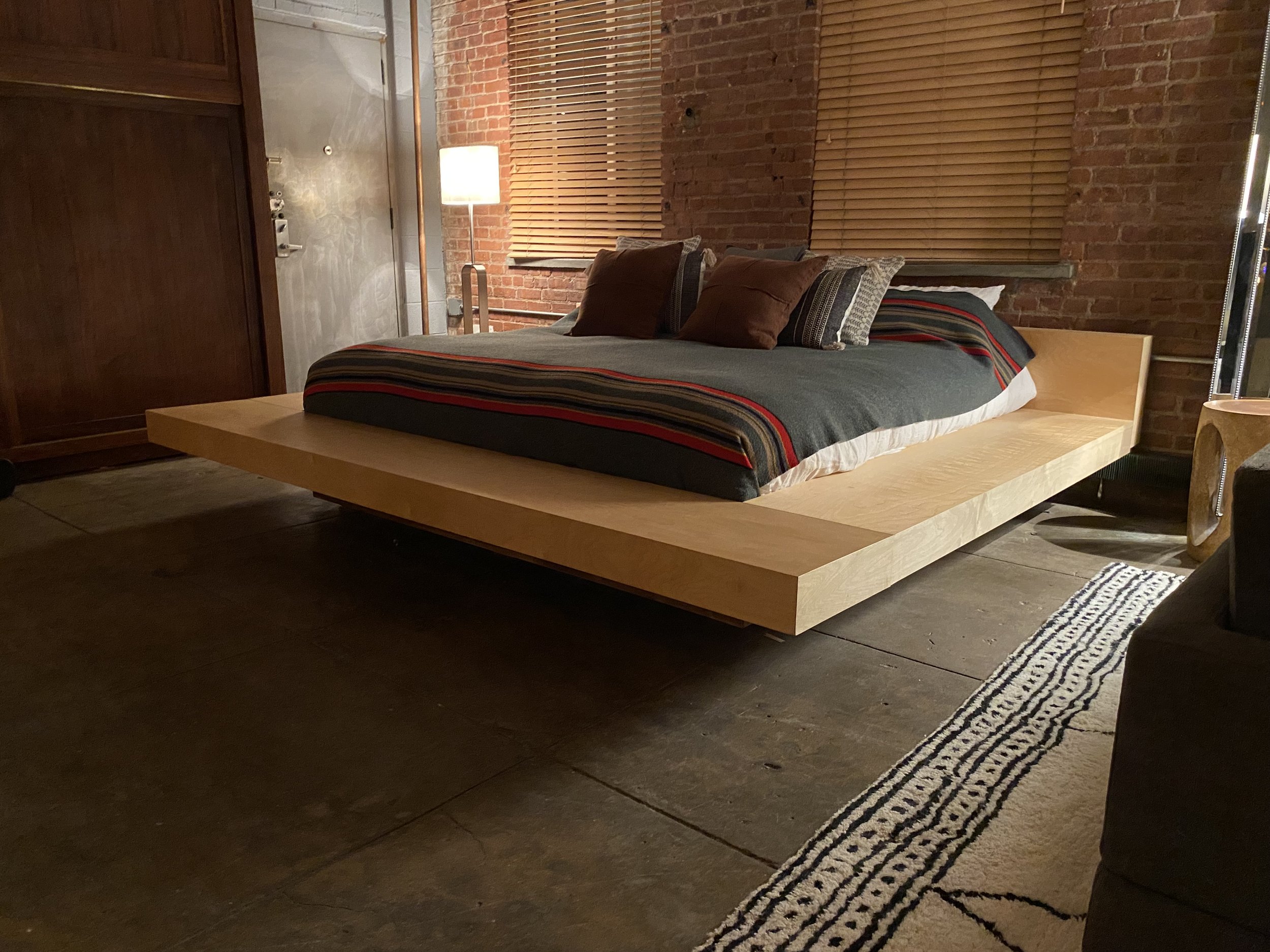 A modern bedroom with a floating wooden bed on a concrete floor, brick walls, a window with wooden blinds, a floor lamp, and side table.