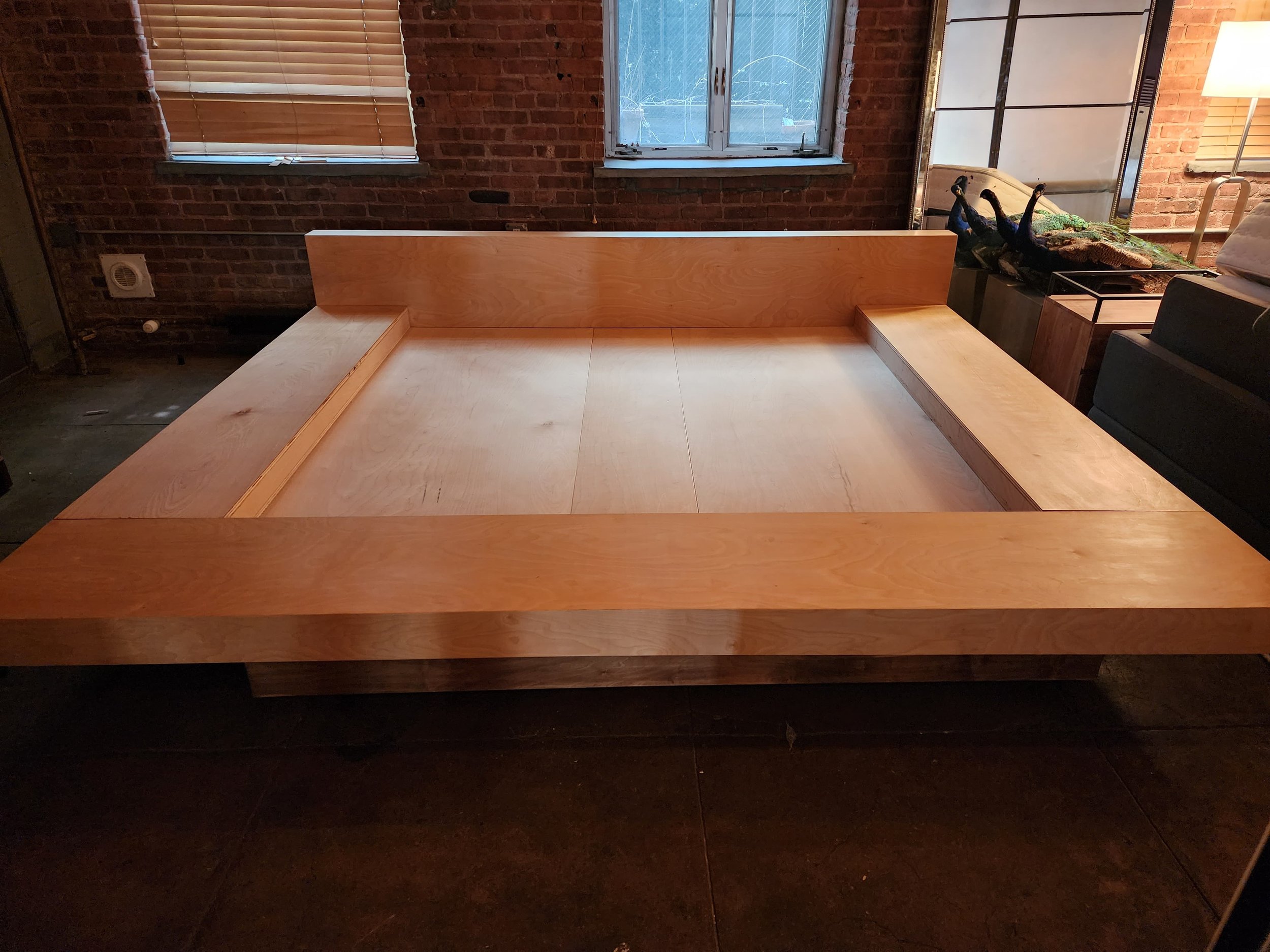 The image shows an unfinished wooden bed frame with a headboard and side rails, set in a room with brick walls, a window with blinds, and a dark floor. There is some furniture and decoration around the room, including a lamp and a sculpture of ducks.
