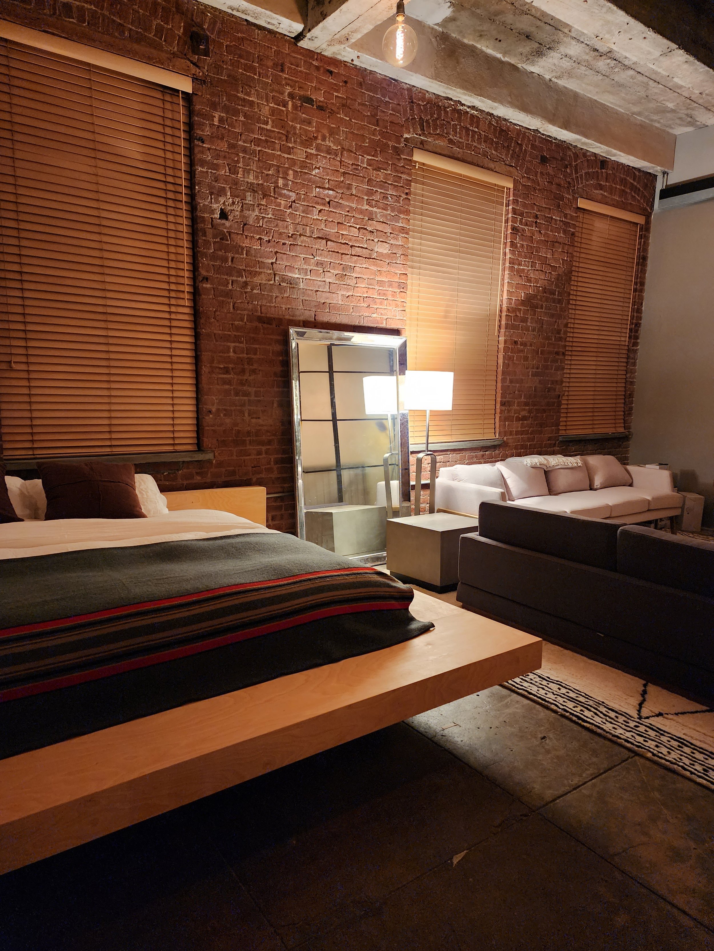 Hotel bedroom with a brick wall, three windows with blinds, a bed with striped blanket, a full-length mirror, a sofa, a small side table, and a floor lamp.