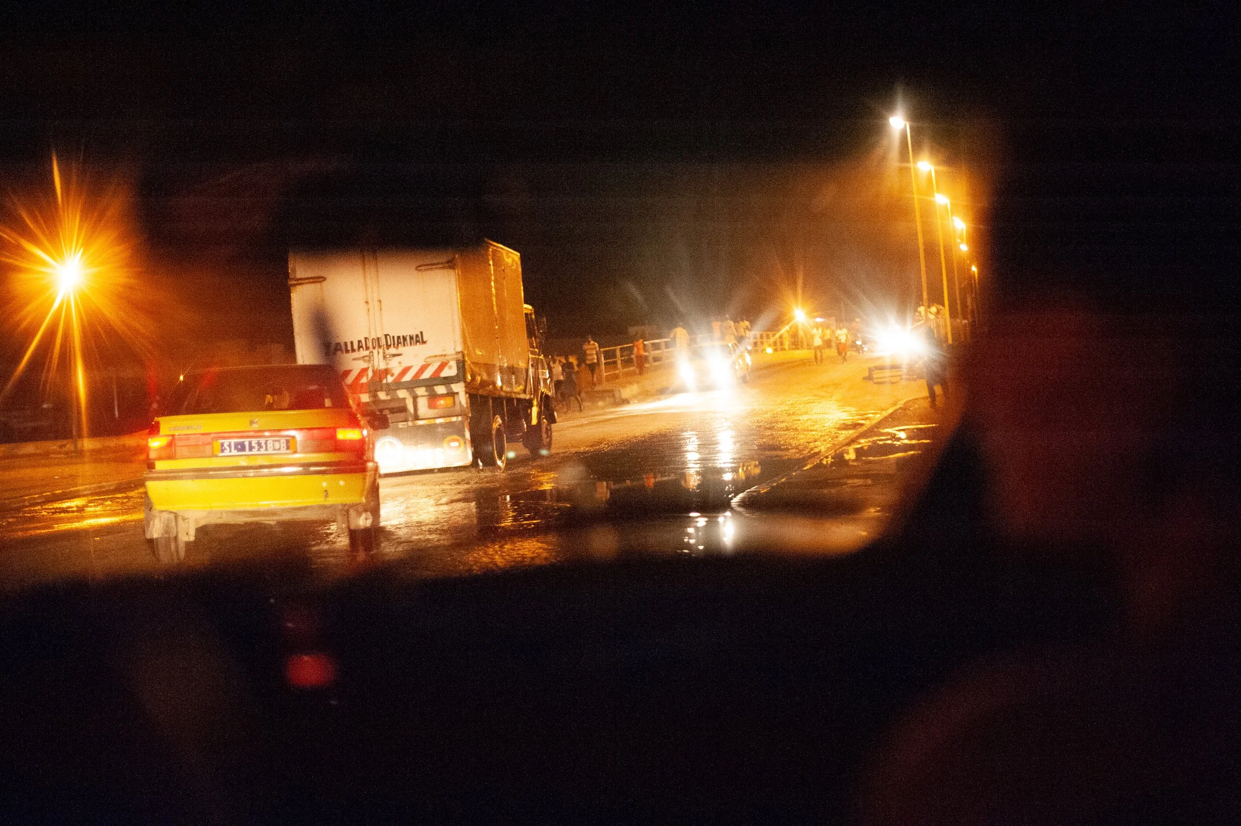 Nighttime road scene with vehicles, including a yellow car and a truck, on a wet and illuminated road.