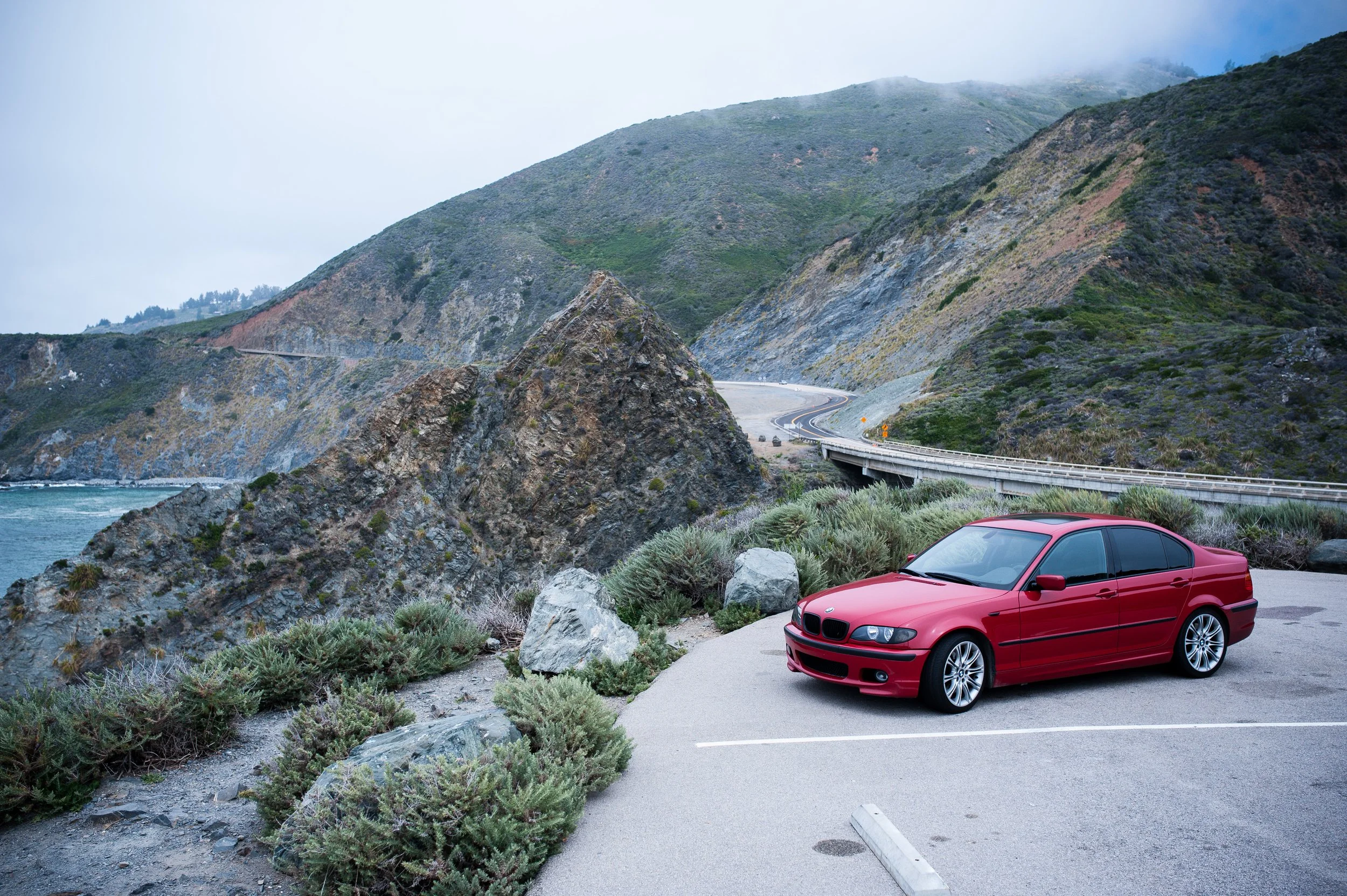 Red car parked near coastal mountain road with rocky landscape and ocean view.
