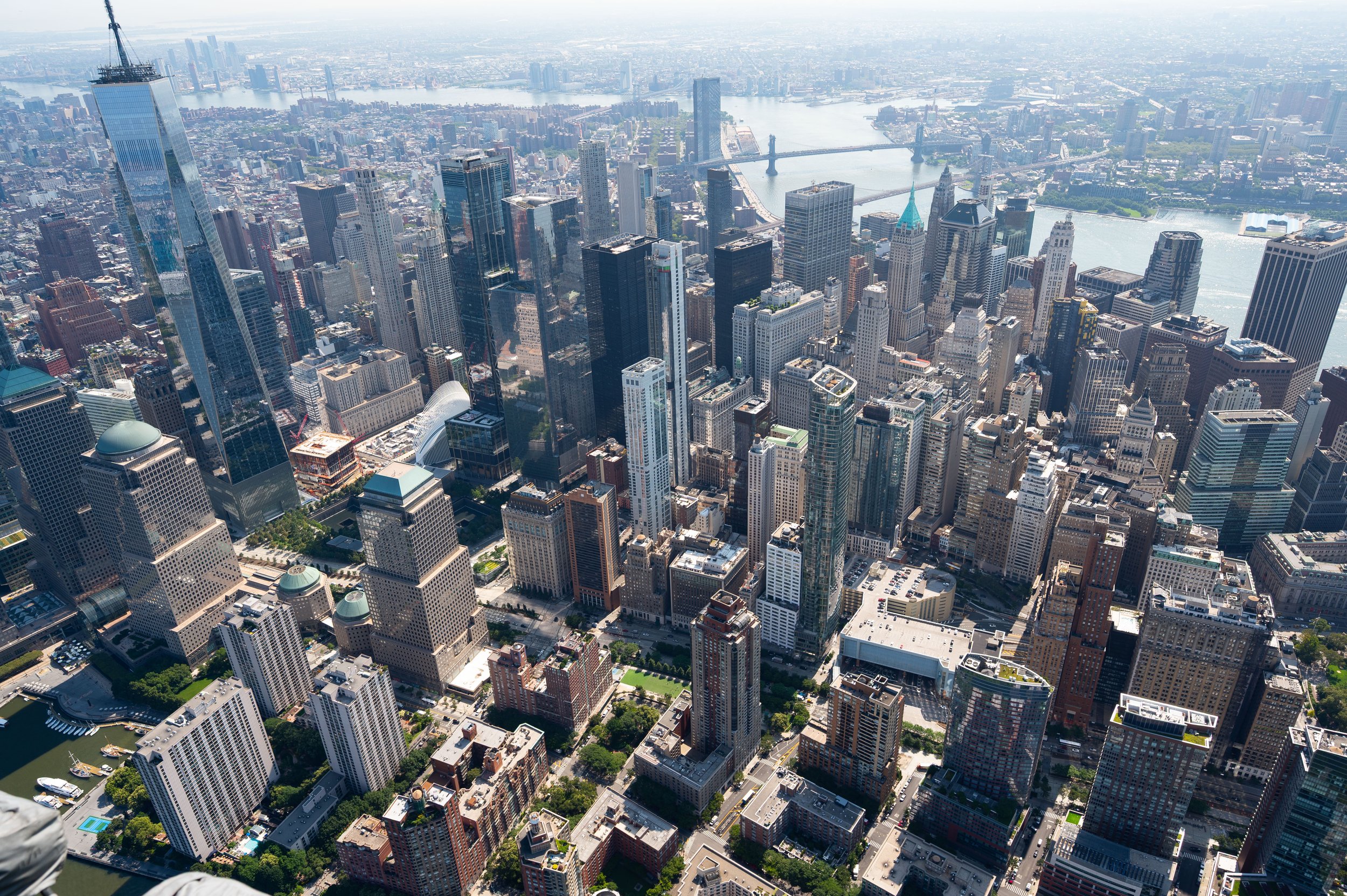 Aerial view of Lower Manhattan featuring One World Trade Center, numerous skyscrapers, bridges, and the surrounding waterfront in New York City.
