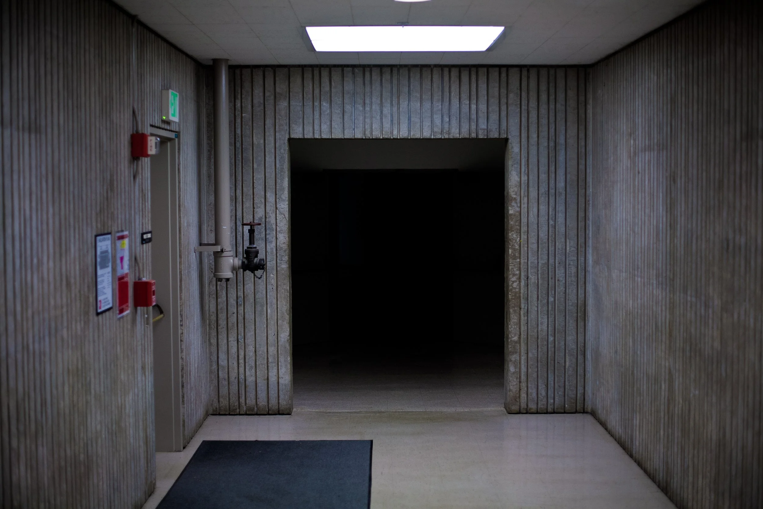 Concrete hallway with dim lighting leading to a dark entrance, featuring industrial-style walls and pipes, exit sign, and emergency fixtures on the side wall.