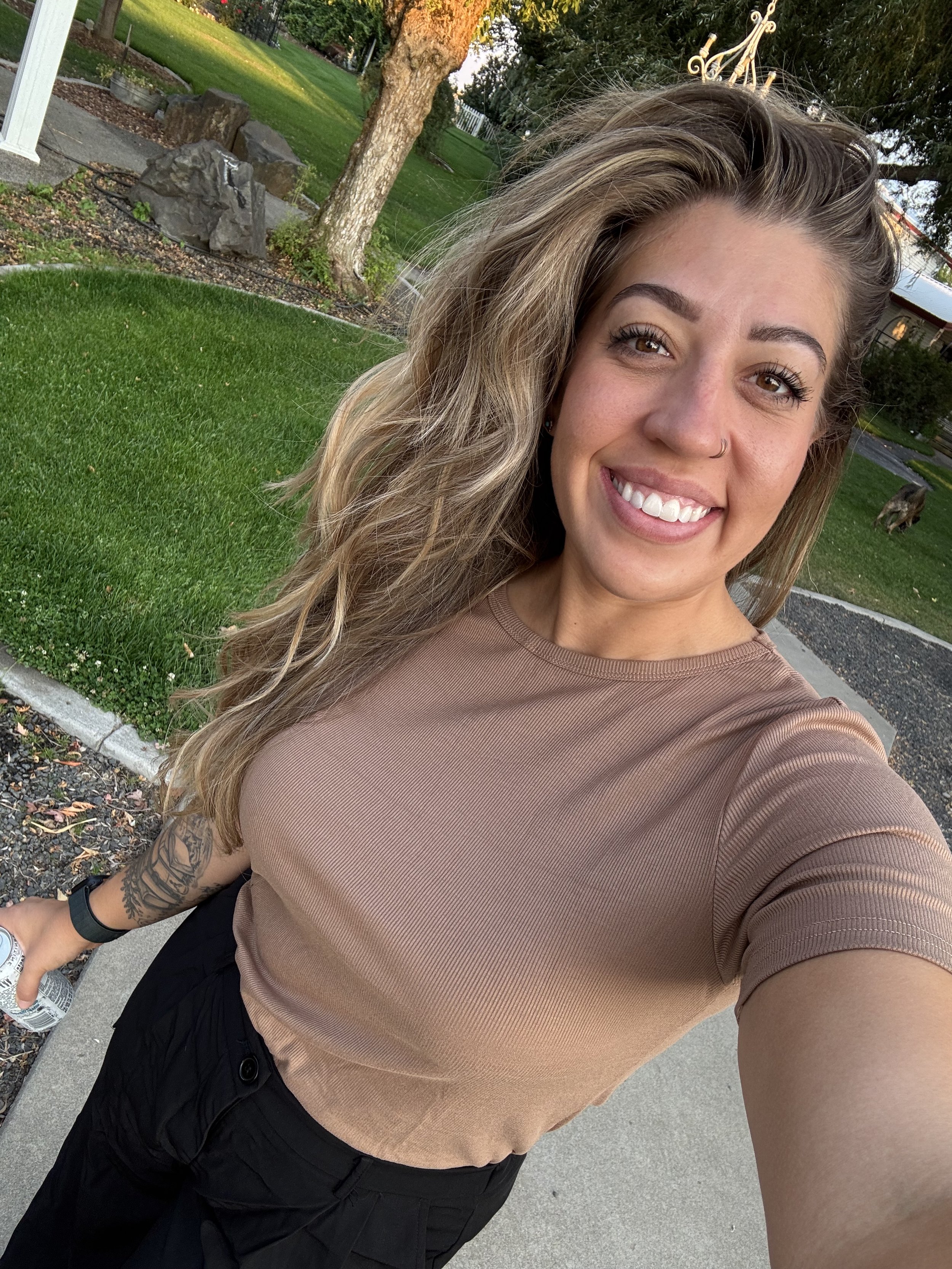 A smiling woman with long wavy hair taking a selfie outdoors in a park-like setting with grass, trees, and rocks in the background.