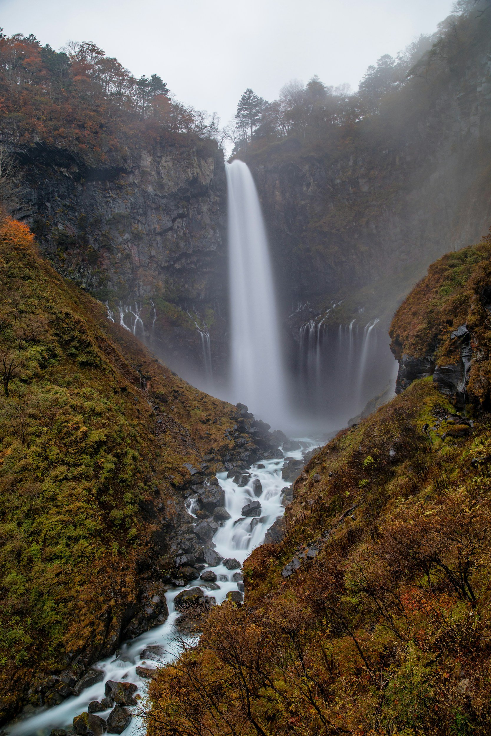 A tall waterfall cascading down a rocky cliff surrounded by autumn-colored trees and flowing into a rocky stream.