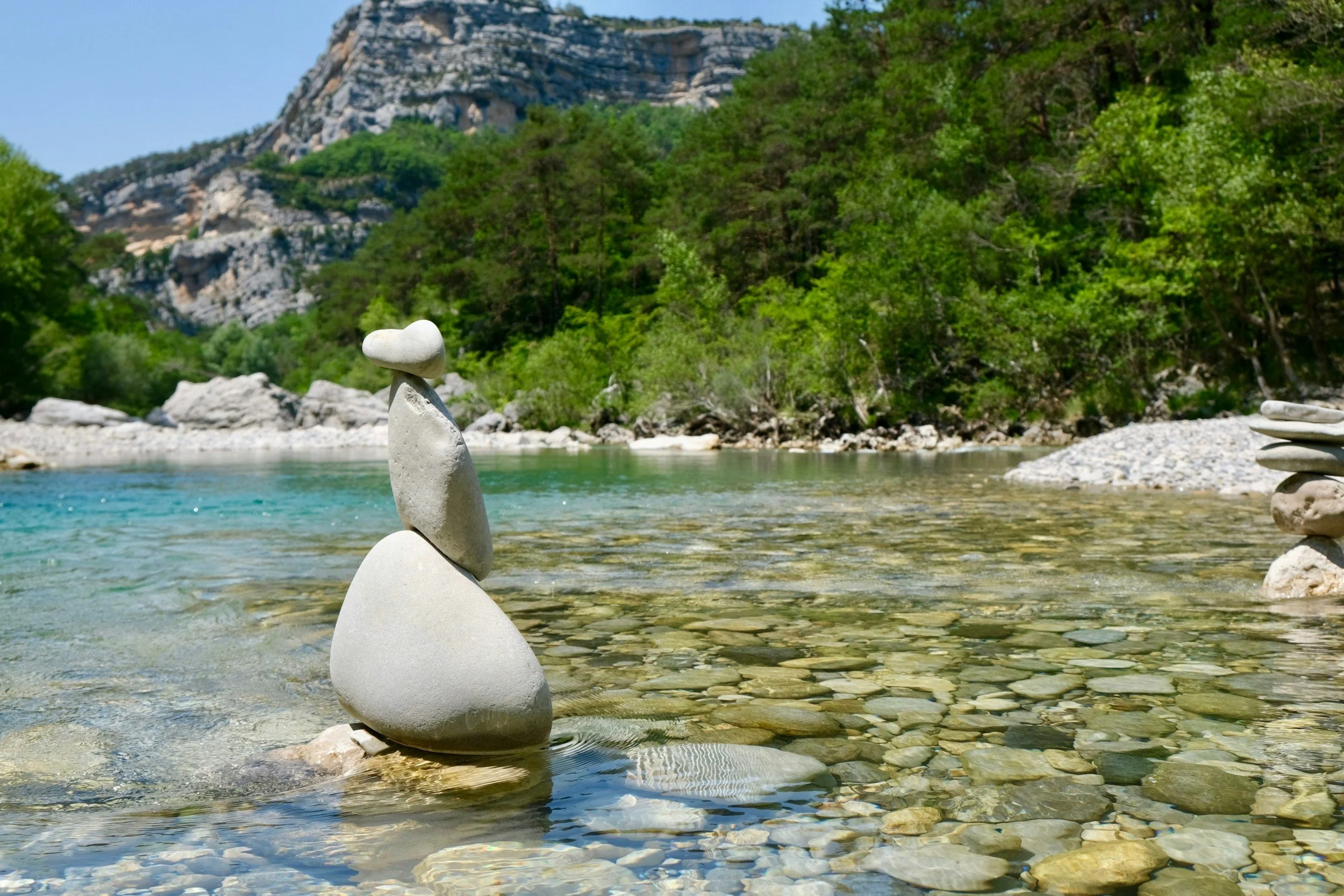 Smooth stones balanced in clear water, backed by green trees and a tall rocky mountain under a bright blue sky — calm and harmonious scene.