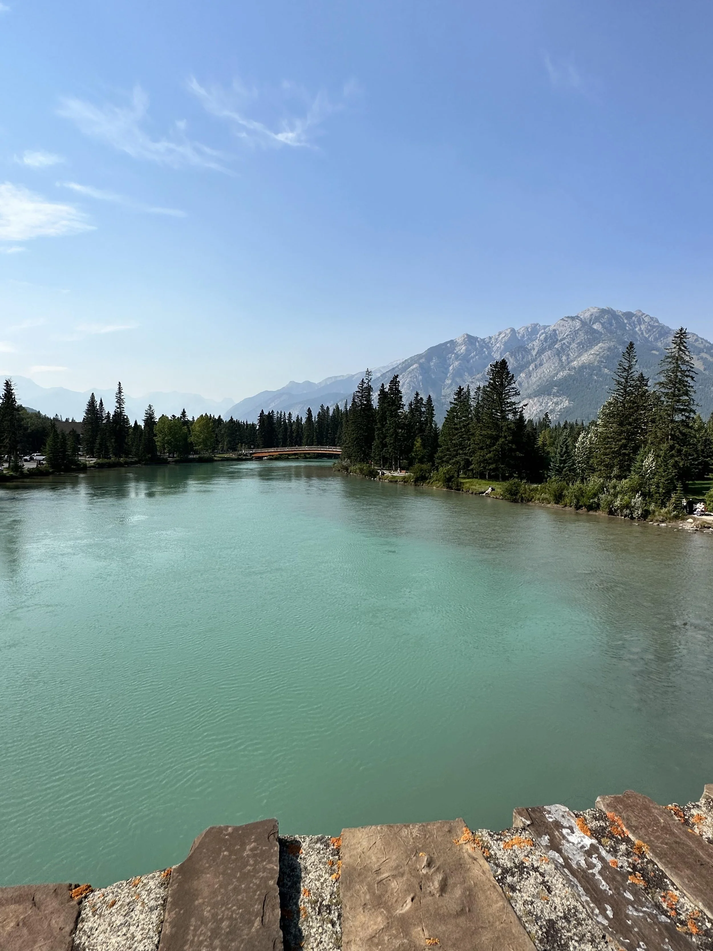 The view from a stone path of a large lake with a border of trees and a mountainous backdrop.